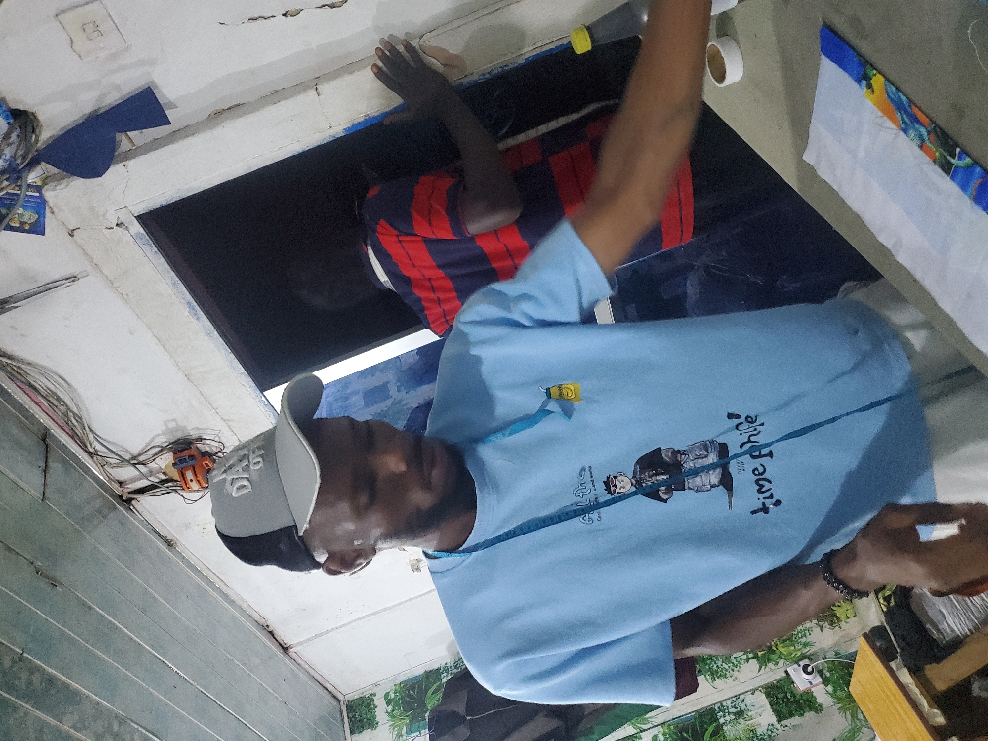 The image shows two men in a dimly lit workshop, likely in Jos, Nigeria. One man, wearing a light blue t-shirt with a graphic of a person and the text "time phife" on it, is the primary subject. He is wearing a grey baseball cap and has a measuring tape draped around his neck. The other man, in the background, is wearing a red and blue striped shirt and appears to be working on something, though his face is obscured.
The workshop appears to be somewhat cluttered, with electrical wires and tools visible. The walls are unpainted and show signs of wear. The lighting is low, suggesting it might be evening or the interior is not well-lit. There is a roll of white tape on a surface behind the main subject. To the right, a piece of fabric with a colorful, possibly abstract pattern is visible.
The overall impression is one of a busy, functional workspace where tailoring or garment production might be taking place. The subjects appear focused on their work.