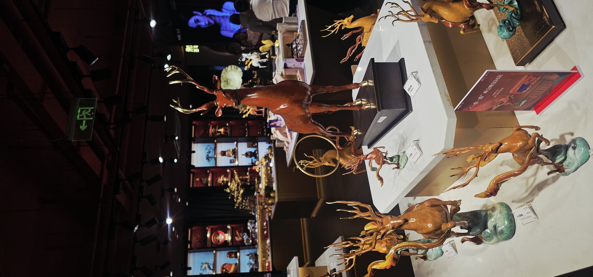 The image depicts the interior of a shop in Yuzhong District, China, featuring a display of numerous bronze deer sculptures. In the foreground, several intricately designed deer figurines are arranged on white display tables. These sculptures vary in pose and detail, with some appearing dynamic and others more static. One prominent sculpture in the center foreground is a large, reddish-brown deer with gold antlers, standing on a dark rectangular base. Behind the foreground displays, more shelves and display cases are visible, filled with various decorative items and smaller sculptures, including what appears to be a golden tree. In the background, shelves are illuminated with spotlights, showcasing a variety of artifacts, some of which seem to be traditional Chinese art pieces. A large screen displays an image of a woman. Several people are visible in the background, browsing or interacting within the shop, although their faces are not clearly discernible. An illuminated green exit sign with an arrow pointing to the left is visible hanging from the ceiling. The lighting suggests an indoor retail environment.