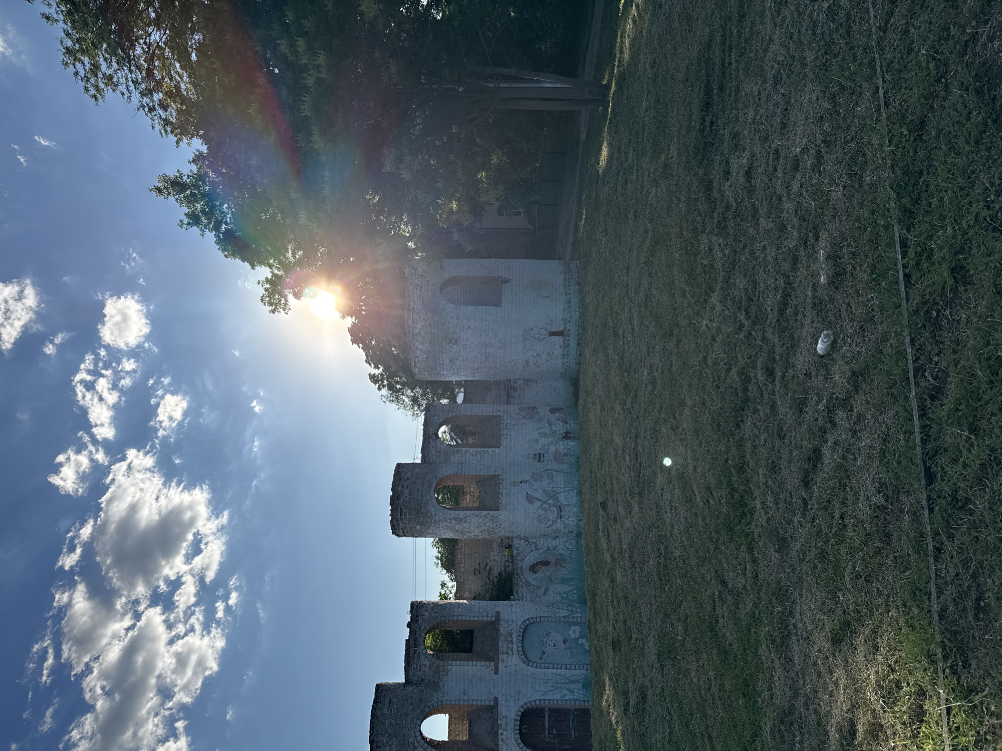This media file is a photograph of the ruins of a white brick structure, possibly an old building or tower, set against a bright blue sky with scattered white clouds. The sun is shining intensely, casting a lens flare and rainbow effect across the upper left portion of the image, suggesting it is midday.
The structure appears to be weathered and in disrepair, with arched openings and sections of missing walls. Faint murals or paintings are visible on some of the brick surfaces, depicting stylized figures and floral elements. Overgrown grass covers the ground around the ruins, with a few sparse trees providing some shade. A single white plastic bottle is visible in the grass in the lower right quadrant of the image.
The overall scene evokes a sense of decay and neglect, yet the bright sunlight and clear sky lend a feeling of warmth and perhaps a hint of melancholy. The image is oriented vertically, with the sky occupying the left side and the ground and structure on the right. There is no visible text in the image.