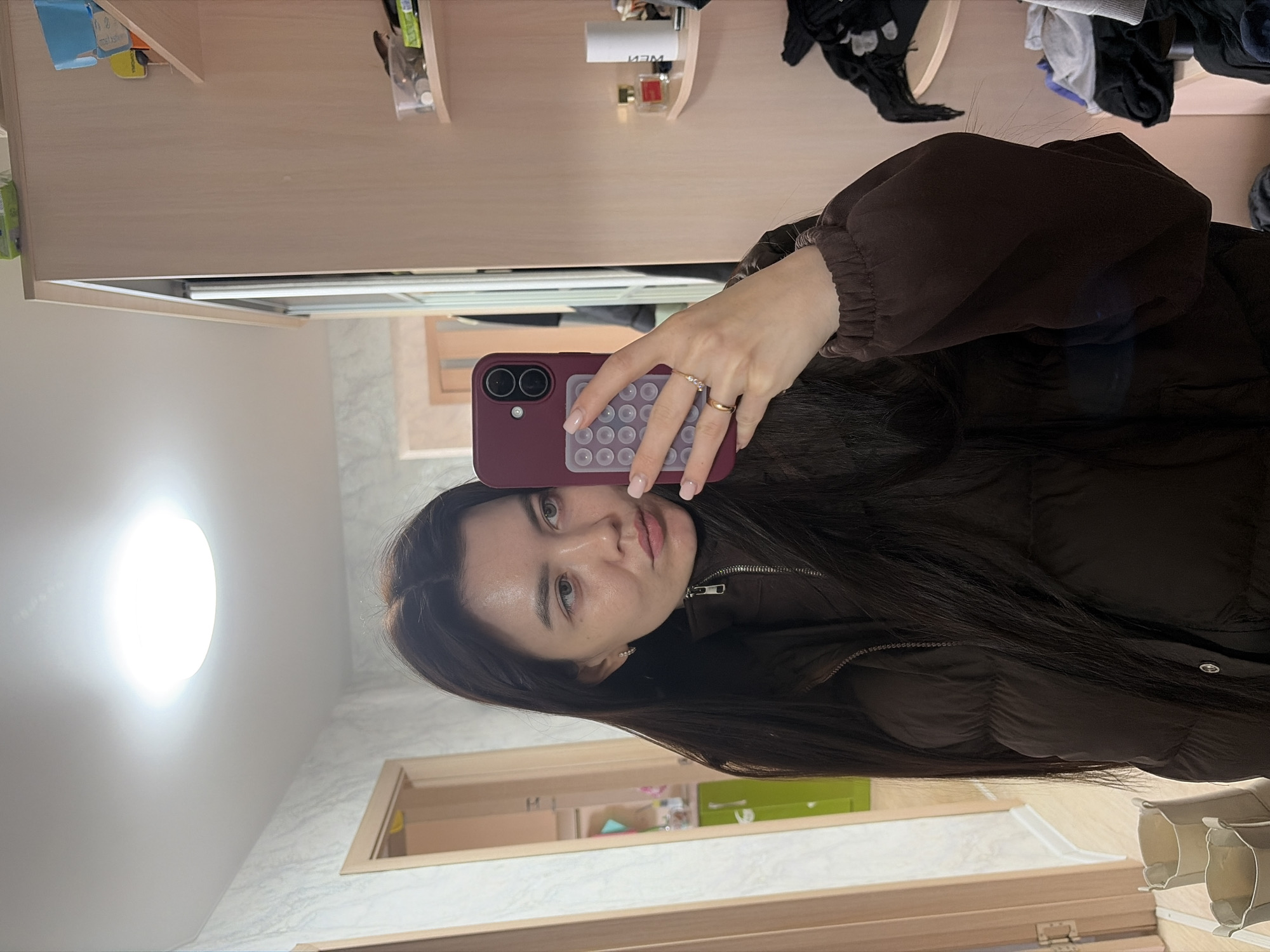 This is a mirror selfie featuring a young woman indoors in Chaikovsky, Russia. The woman, with long, dark hair and light-colored eyes, looks directly at the camera with a neutral expression. She is wearing a dark brown, possibly puffy or quilted jacket with elasticated cuffs and a zippered front. On her left hand, she wears two rings: one thin gold-colored ring with small stones, and a plain gold-colored band. She holds a smartphone with a dark red/burgundy case that has a light-colored, circular multi-bumped grip or pop-socket on its back.
The background, visible in the mirror reflection, depicts an indoor setting with light-colored wooden shelves or cabinets. These shelves hold various items, including a blue box, clear containers with small objects, and perfume bottles. A white container with the Cyrillic text "NЭИ" or "ИЭМ" (NEM or IEM) is also visible. Nearby, some folded clothes in dark, gray, and blue tones are stacked. A bright circular light fixture on the ceiling illuminates the room. The walls appear to have a light-colored, subtly patterned texture, possibly wallpaper or tiles, and a wooden door frame is also visible. The presence of artificial lighting suggests an indoor setting where the time of day is not clearly indicated.