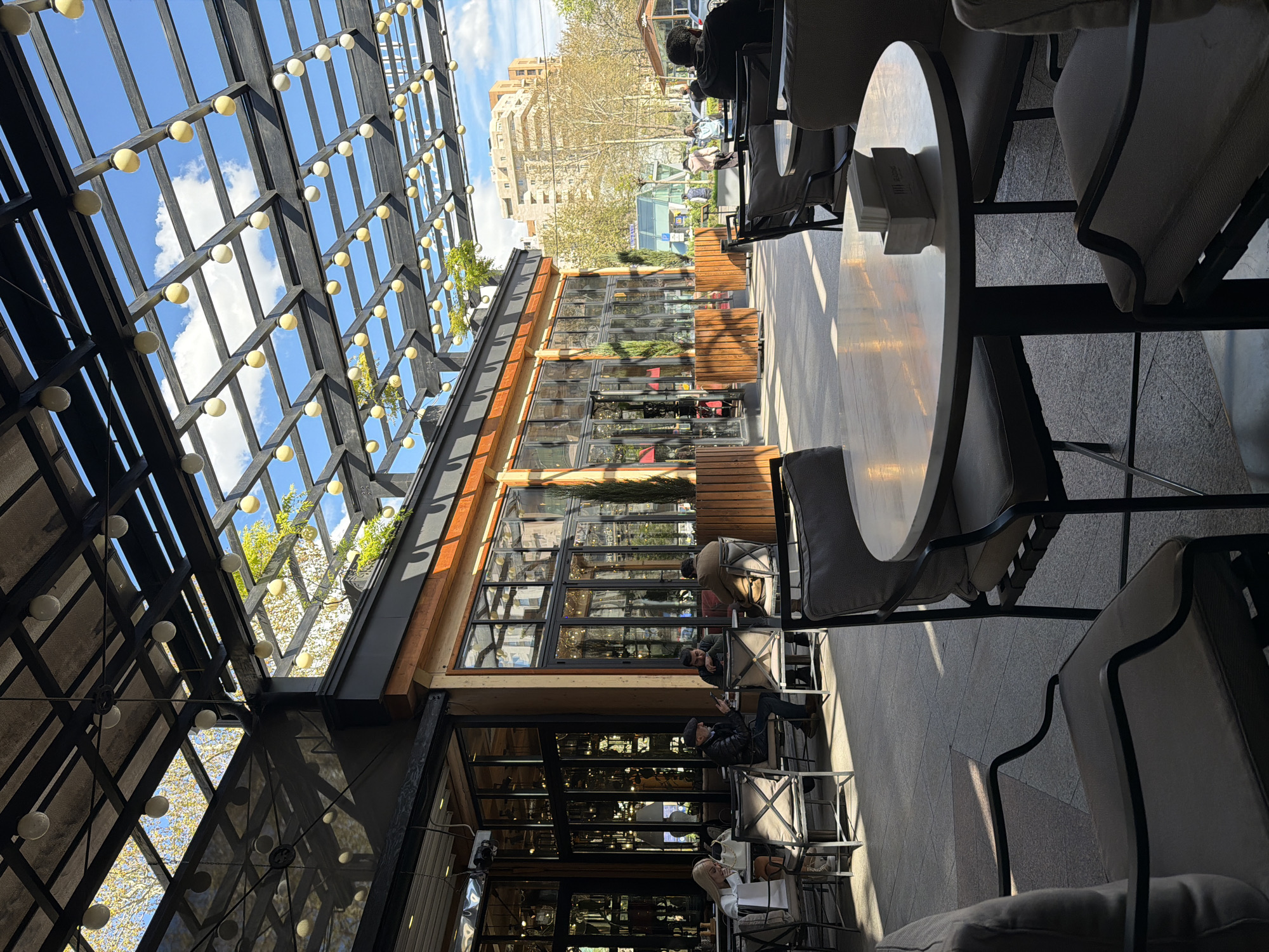 The image shows an outdoor patio space in Yerevan, Armenia, possibly a café or restaurant. The patio is covered by a black metal frame structure with evenly spaced, spherical lights. Through the open structure, a blue sky with scattered white clouds is visible.
Adjacent to the patio is a building with a facade composed of large glass windows and wooden accents. Inside the building, people are seated.
The patio itself contains round tables with grey cushioned chairs. People are seated at some of the tables, engaged in activities such as reading or using their phones.
Outside the patio, across a pedestrian area, a multi-story building can be seen. Trees are scattered in the background, suggesting an urban setting.