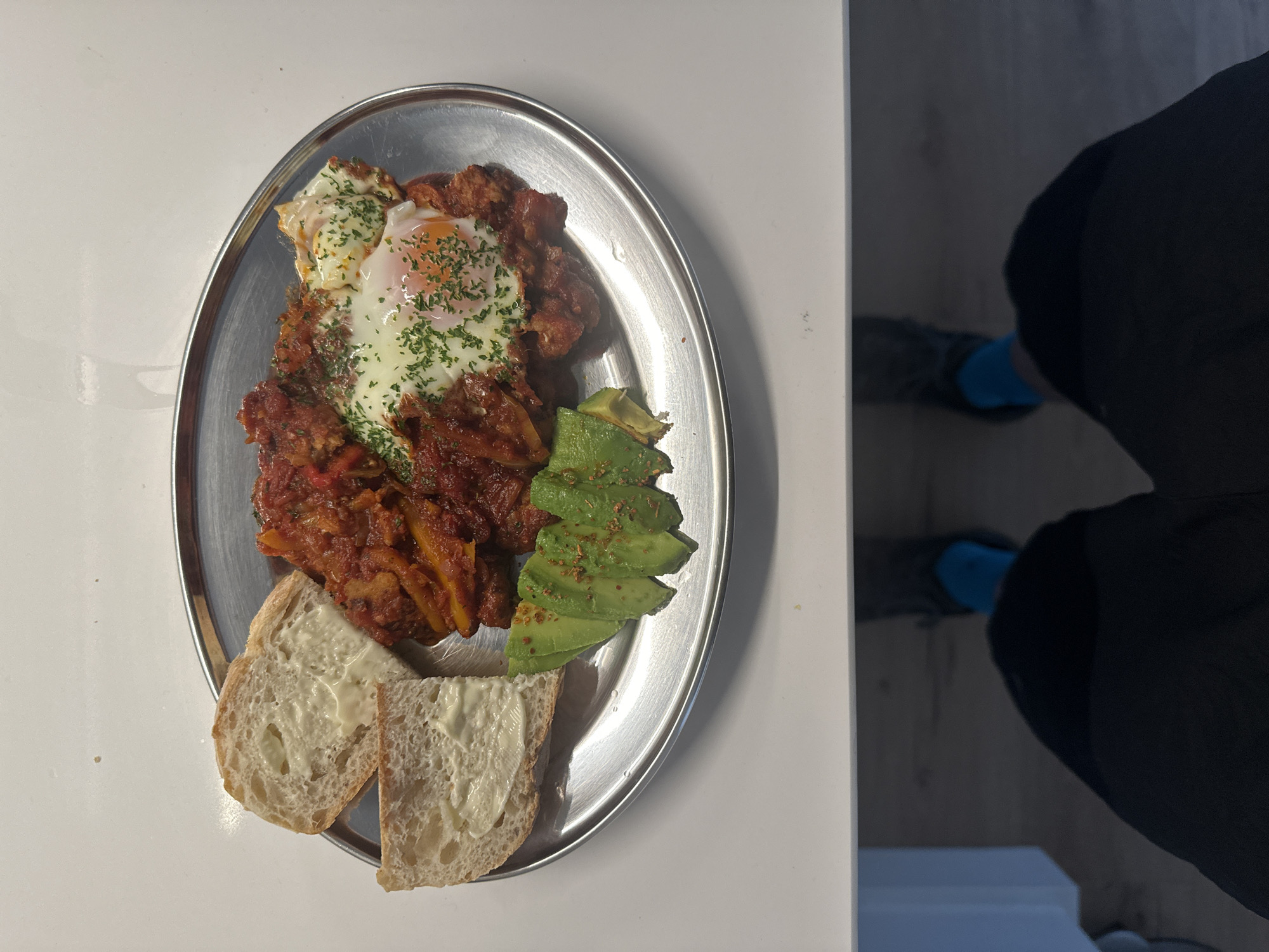 A top-down shot shows a silver oval platter of food and a person in black clothing. The platter contains two fried eggs topped with green herbs, a tomato-based stew with yellow peppers, slices of avocado with spices, and two slices of buttered bread. The platter sits on a white surface next to a dark reflection. Part of a person is visible to the right, wearing black pants and grey shoes with blue socks. The setting appears to be indoors, possibly in a kitchen or dining area.