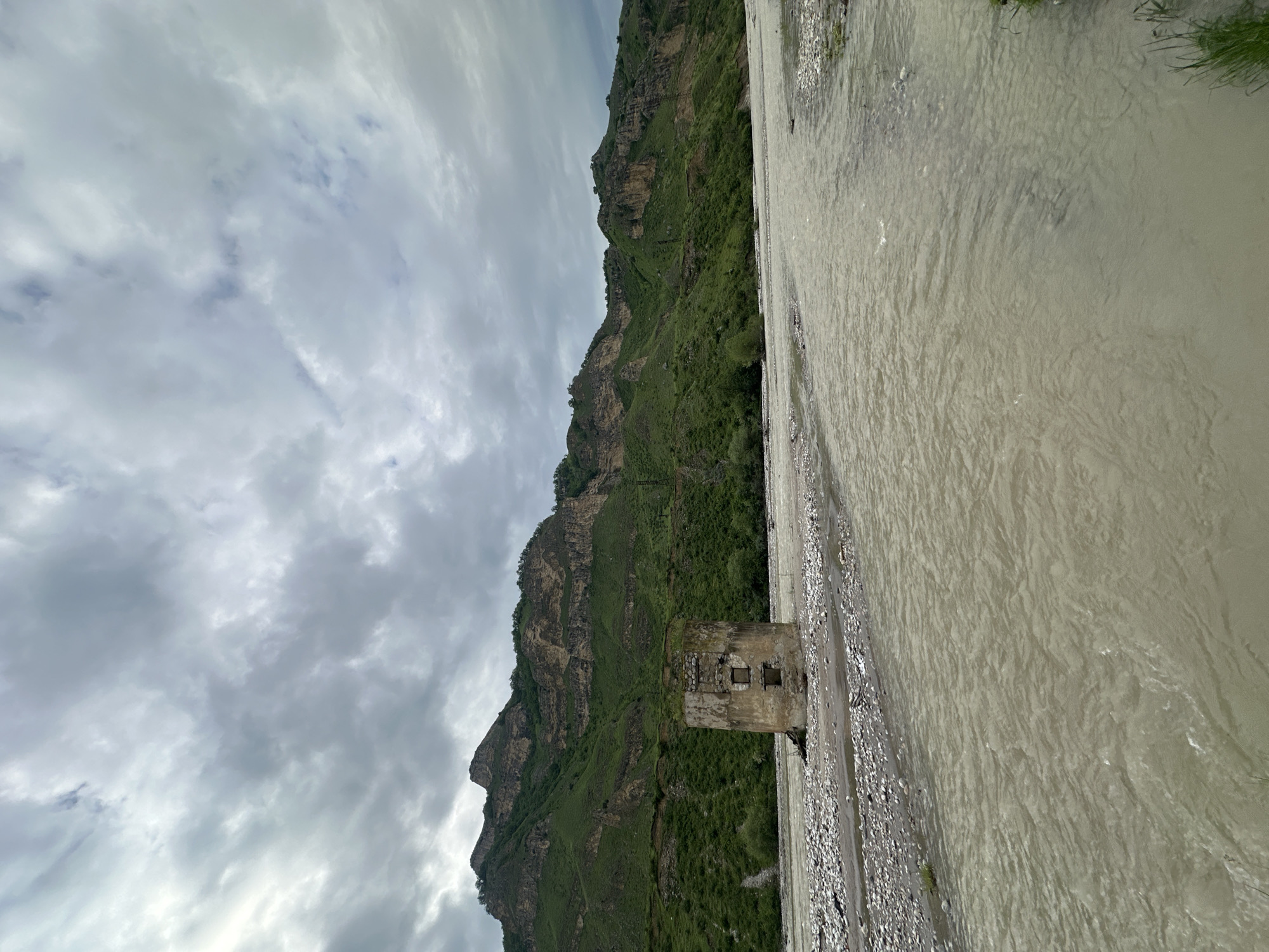 In the city of Bedyk, Russia, a weathered, cylindrical stone structure stands partially submerged in a wide, murky river. The structure appears to be a ruin, with visible square openings on its front facade. The riverbed is strewn with smooth, grey pebbles. In the background, a lush green mountain range rises, its slopes covered in vegetation and crisscrossed by what appear to be ancient terraces or natural rock formations. The sky above is overcast with thick, grey clouds.