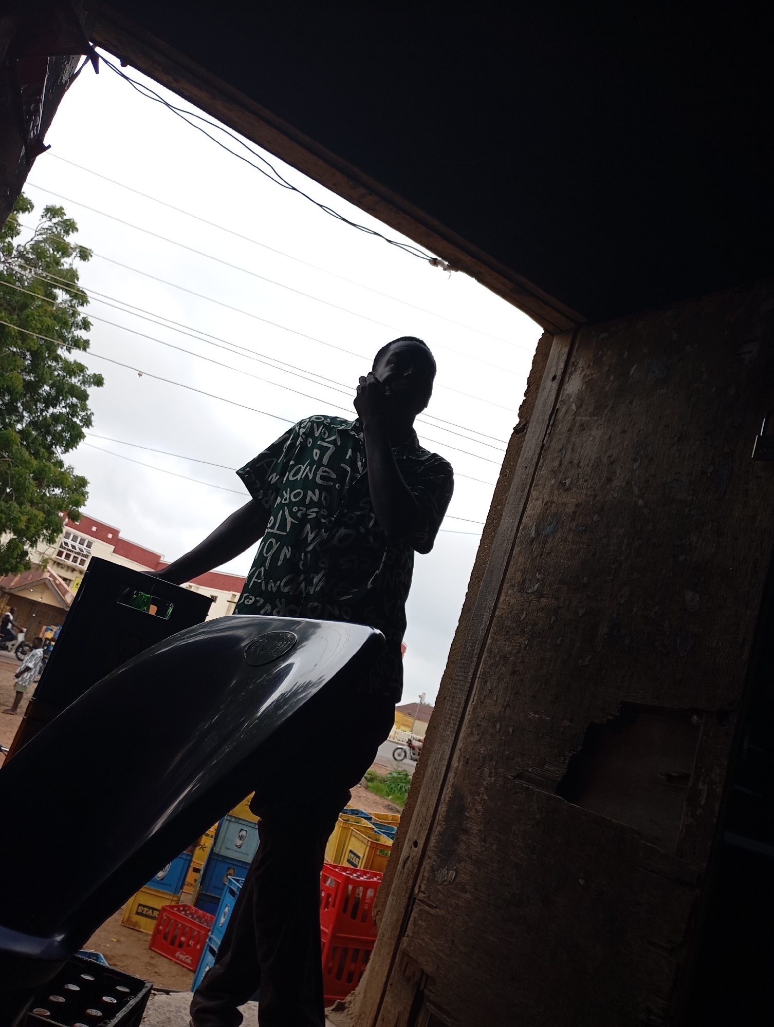 A low-angle, full-shot image captures a man standing in what appears to be an open doorway or shop front in Shendam, Nigeria. The man is silhouetted against a bright sky and is wearing a dark, patterned shirt. He is holding a black object, possibly a car part or a large container, with his left hand. His right hand is raised to his face, obscuring it. In the foreground, to the left of the man, is the shiny, dark surface of another object, also possibly a car part. Behind the man, a stack of colorful crates, predominantly red, yellow, and blue, are visible. One crate has "STAR" written on it. The background shows a building with a red roof and a tree with green foliage. Power lines are also visible crisscrossing the sky. The overall impression is of a market or a commercial area.