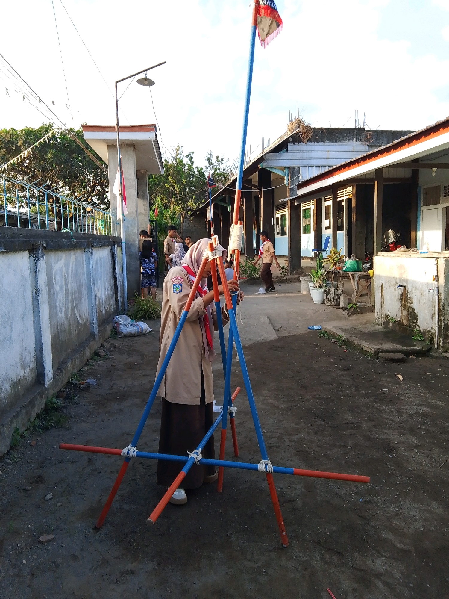 In Kuripan Utara, Indonesia, a young person wearing a brown scout uniform and a headscarf stands in a dirt courtyard, focused on constructing a tripod-like structure with blue and orange poles. The structure is held together with white rope. In the background, other children in scout uniforms and civilian clothing mill about near buildings with open doorways and windows. A flag is visible on a tall pole. The environment appears to be a school or community gathering area, with a concrete wall and some trees. The lighting suggests it is late afternoon.