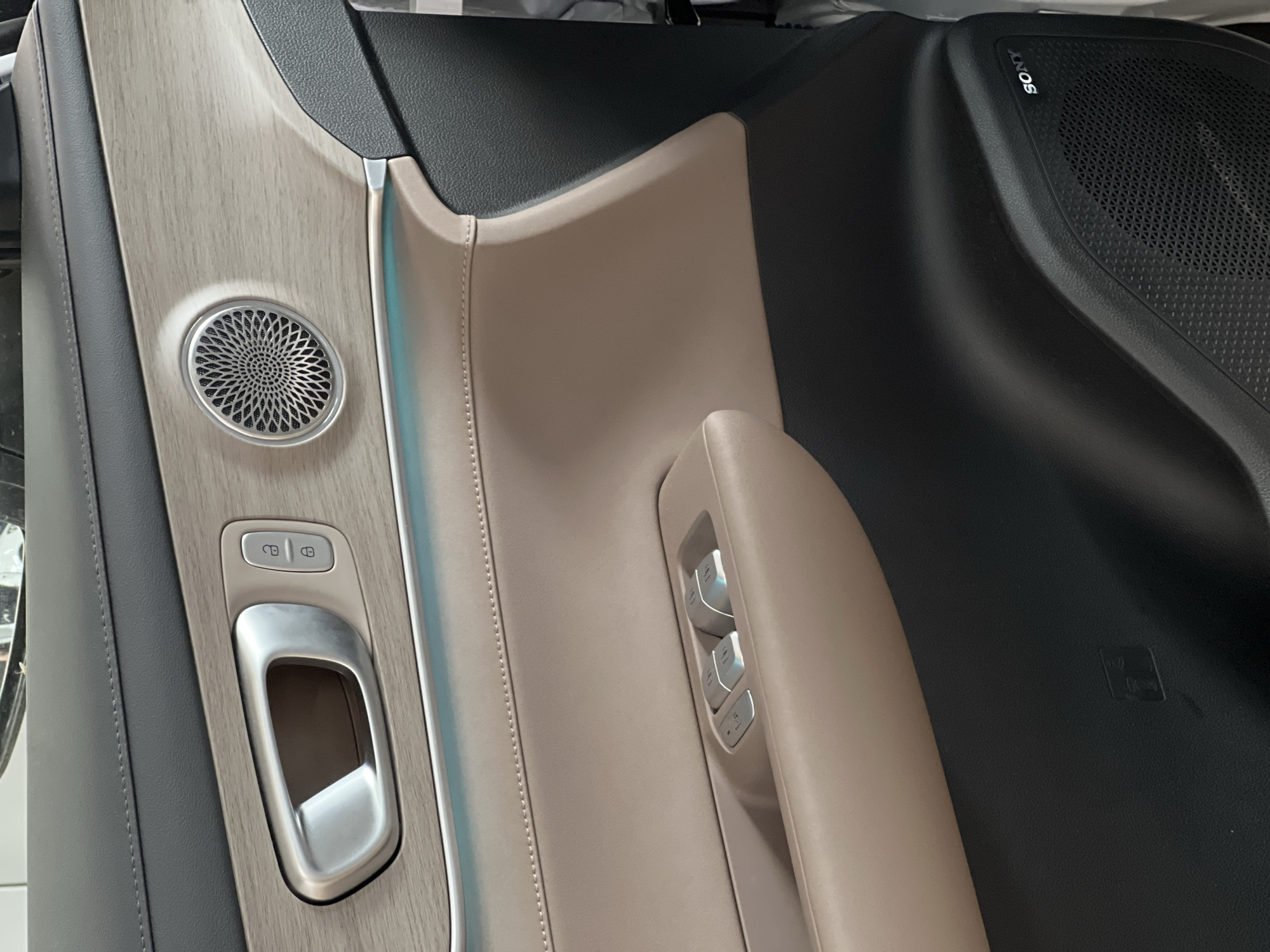 This is a close-up interior shot of a car door, likely a modern vehicle due to the materials and design. The door panel features a combination of materials: a dark gray textured upper section, a wood-grain accent panel, and a light tan leather lower section. Prominently visible is a brushed metal interior door handle, flanked by lock and unlock buttons on the wood-grain panel. Below the wood-grain panel, a thin strip of ambient blue light runs across the width of the door. The tan leather section houses the window control panel, with several silver buttons. Further down, the lower portion of the door is in dark gray plastic, with a visible "SONY" logo indicating a speaker. The image appears to be taken indoors, possibly in a car dealership or private garage, given the lighting and lack of outdoor context. There is no visible indication of the specific location within the city of Izhevsk, Russia; the image solely depicts the interior of a vehicle.