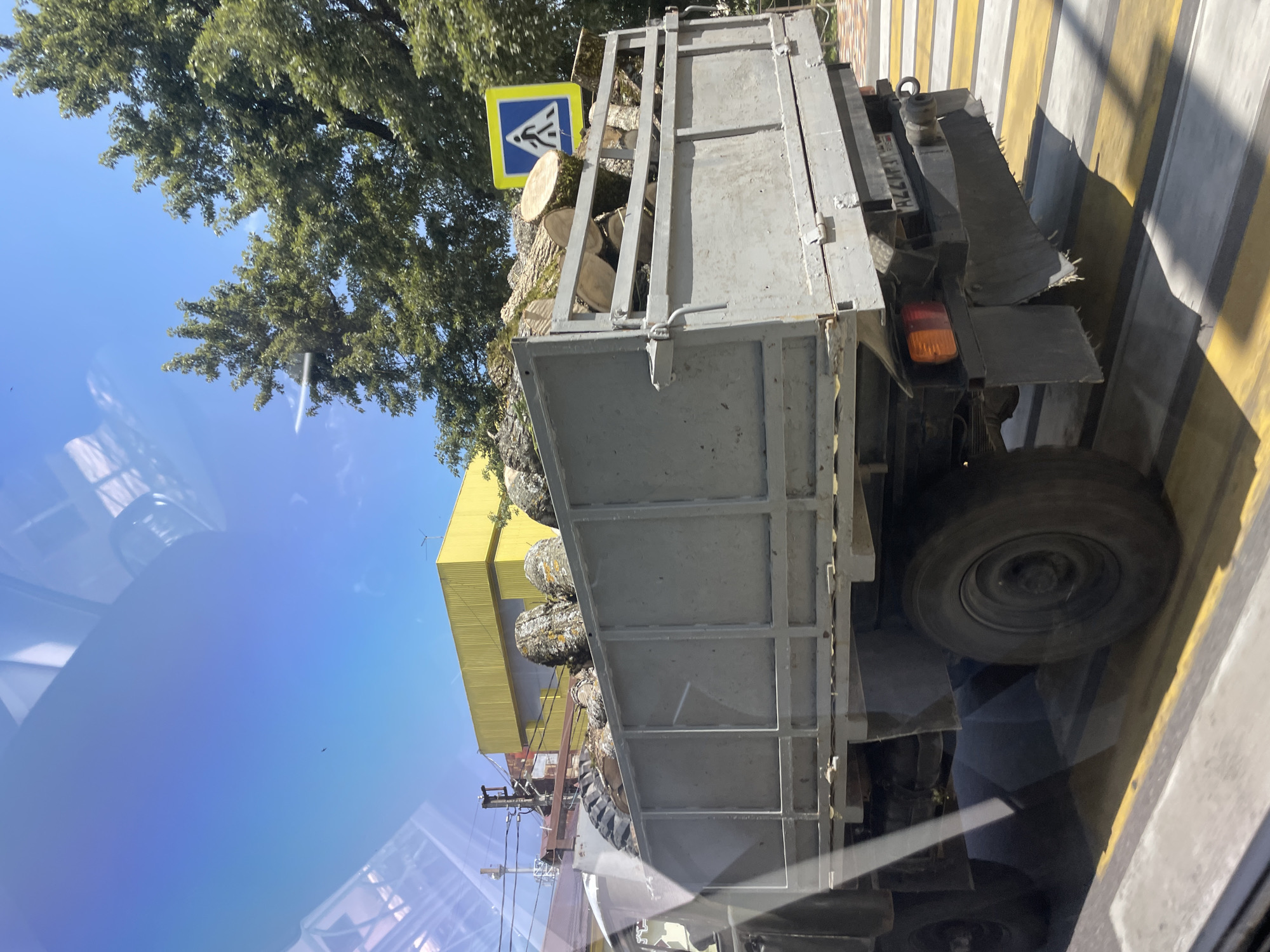 The image shows a grey flatbed truck with its bed filled with logs. The truck is driving across a pedestrian crossing with yellow and white stripes. In the background, there is a yellow building with a ribbed facade, and some green trees. A blue and yellow pedestrian crossing sign is visible on the right side of the frame. The sky is clear and blue with some reflections on what appears to be a window. The scene is set in a city environment, likely Stavropol, Russia, given the context.