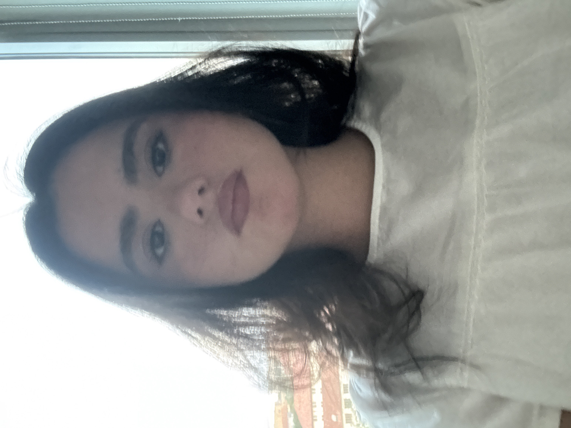 A young woman with dark hair is photographed from the chest up against a bright, blown-out background. She is looking directly at the camera with a neutral expression. Her hair is dark and appears wavy, framing her face. She is wearing a white top with subtle textured detailing. The background is largely white, suggesting a brightly lit environment, possibly near a window. In the lower left portion of the image, out of focus, are hints of buildings with red roofs, suggesting an urban setting. The overall impression is of a casual, indoor portrait. The image does not provide any information to indicate the location is in Austin, United States of America.