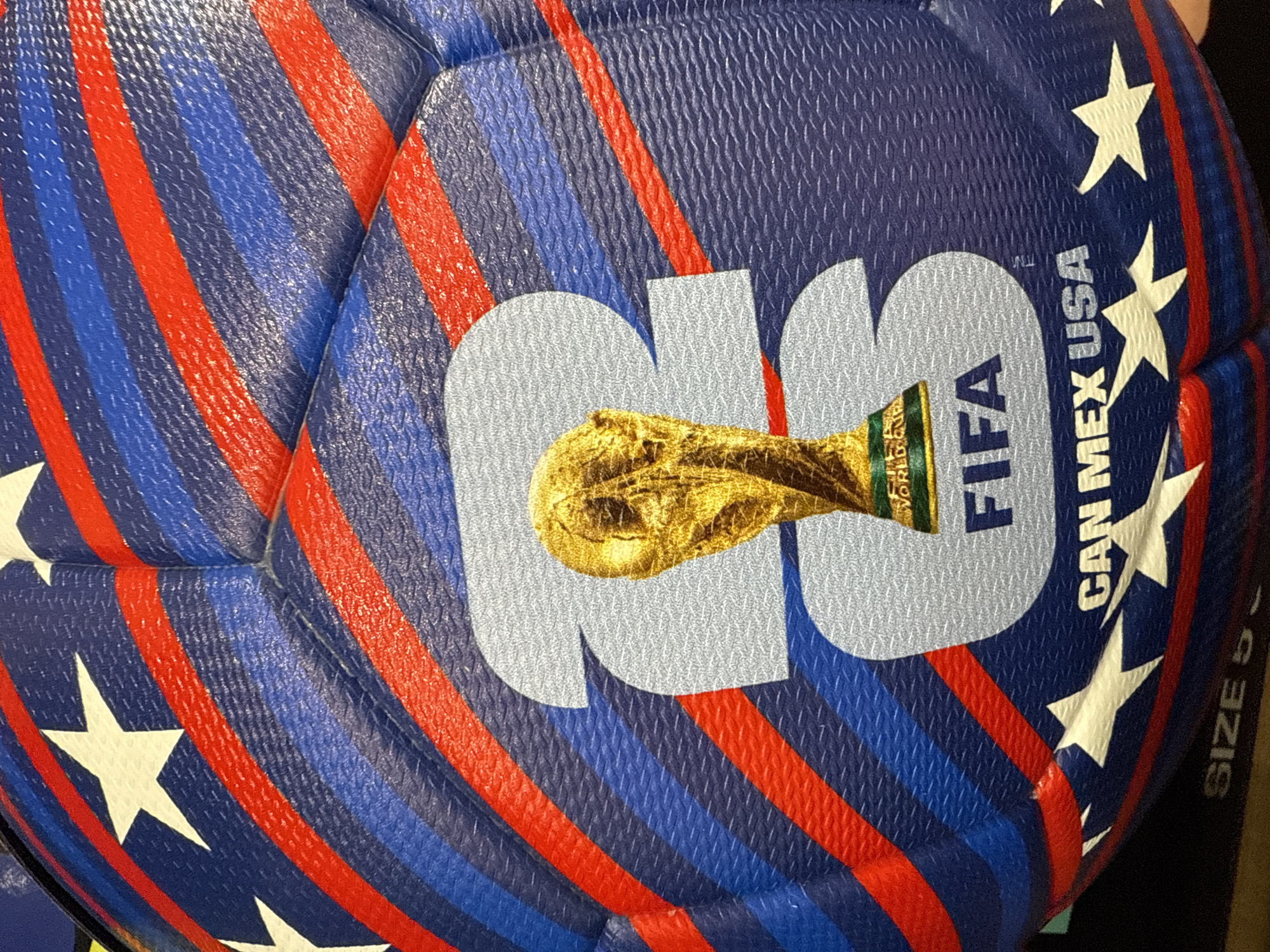 The image shows a close-up of a soccer ball with a red, white, and blue design reminiscent of the American flag. The ball has a large "26" logo on it, along with the FIFA logo and a stylized image of the World Cup trophy. It also says "CAN MEX USA," which indicates the countries hosting the 2026 FIFA World Cup. The ball has white stars on it. Size 5 is printed on the ball. The photo was taken in Bolingbrook, United States of America.