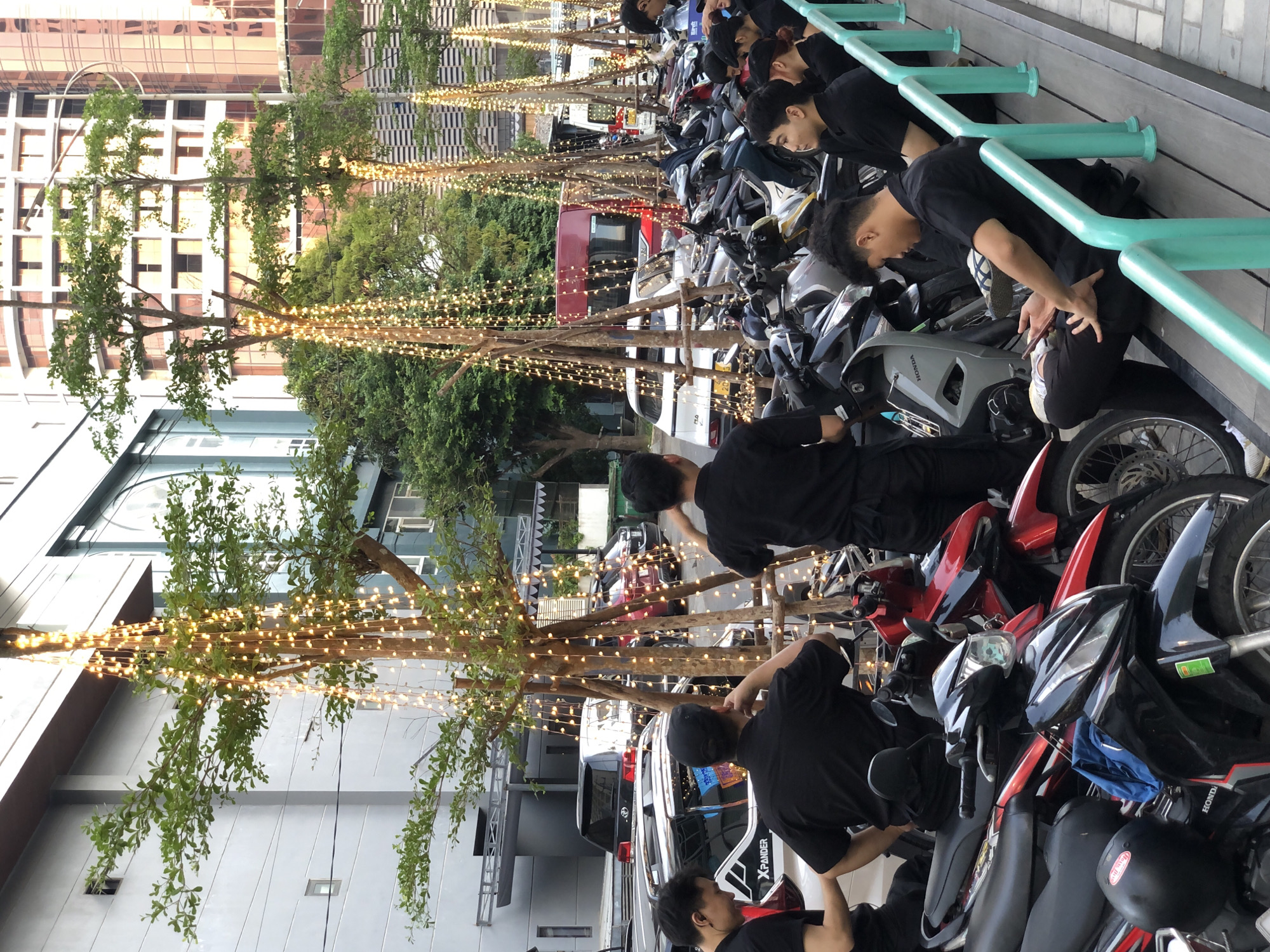 In Đà Nẵng, Vietnam, a group of young men in black shirts and pants are gathered near a row of parked motorcycles. The scene appears to be outdoors, possibly on a sidewalk or pedestrian area adjacent to a street. In the background, several cars and a red bus are visible, along with modern buildings and trees adorned with cascading string lights. The men are engaged in various activities: some are standing, some are sitting on a wooden platform, and one is looking at his phone. The overall atmosphere suggests a casual gathering, possibly during an event or a break.
