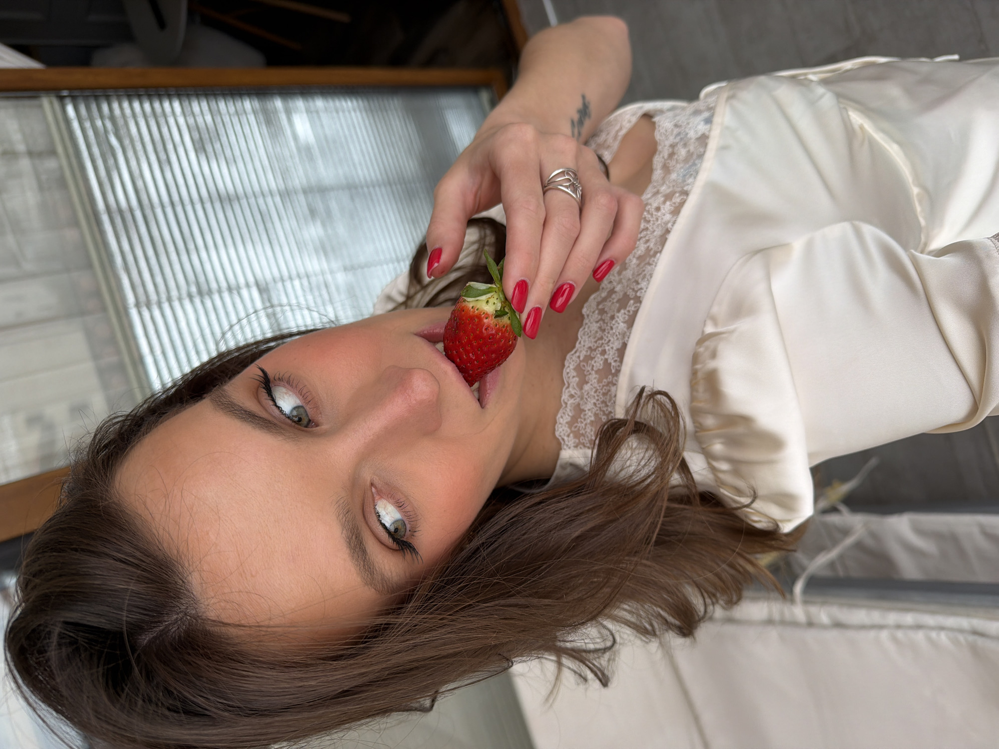 The scene depicts a close-up, slightly elevated perspective of an adult female subject indoors in Budva, Montenegro. The female, with brown hair and green eyes, is positioned facing forward and slightly angled to her right, looking off-camera to her right. She wears a light-colored, possibly cream or ivory, satin-like top with lace trim detailing. Her left hand, adorned with red nail polish and a silver-toned ring on the ring finger, holds a single red strawberry with its green stem intact, which is partially inserted into her mouth. A tattoo is visible on her left forearm. The background is a blurred interior space, revealing a window with horizontal blinds on the right side and a portion of light-colored furniture, possibly a sofa, on the left. A small, white, minimalist table is partially visible in the far right background.