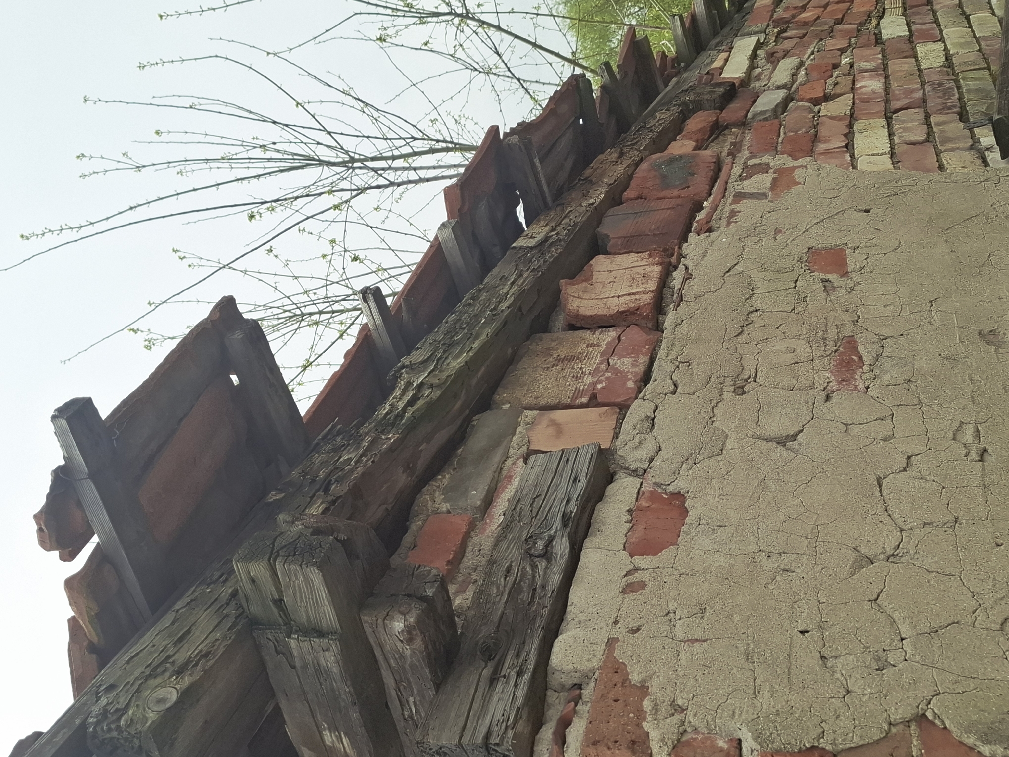 A low-angle shot of a deteriorating building, the location is Unknown, Unknown. The photograph is taken close to the structure, focusing on the intersection of a dilapidated roof and a crumbling exterior wall. The roof, constructed with red clay tiles, is visibly damaged, featuring fractured and displaced tiles. Supporting the roof are weathered wooden beams, some exhibiting signs of decay and rot. The wall is constructed of red bricks, partially covered with a layer of cracked and peeling plaster. The texture of the plaster is rough, with numerous fissures and missing sections exposing the underlying brick. Above the roofline, sparse branches from a tree are visible against a light sky.