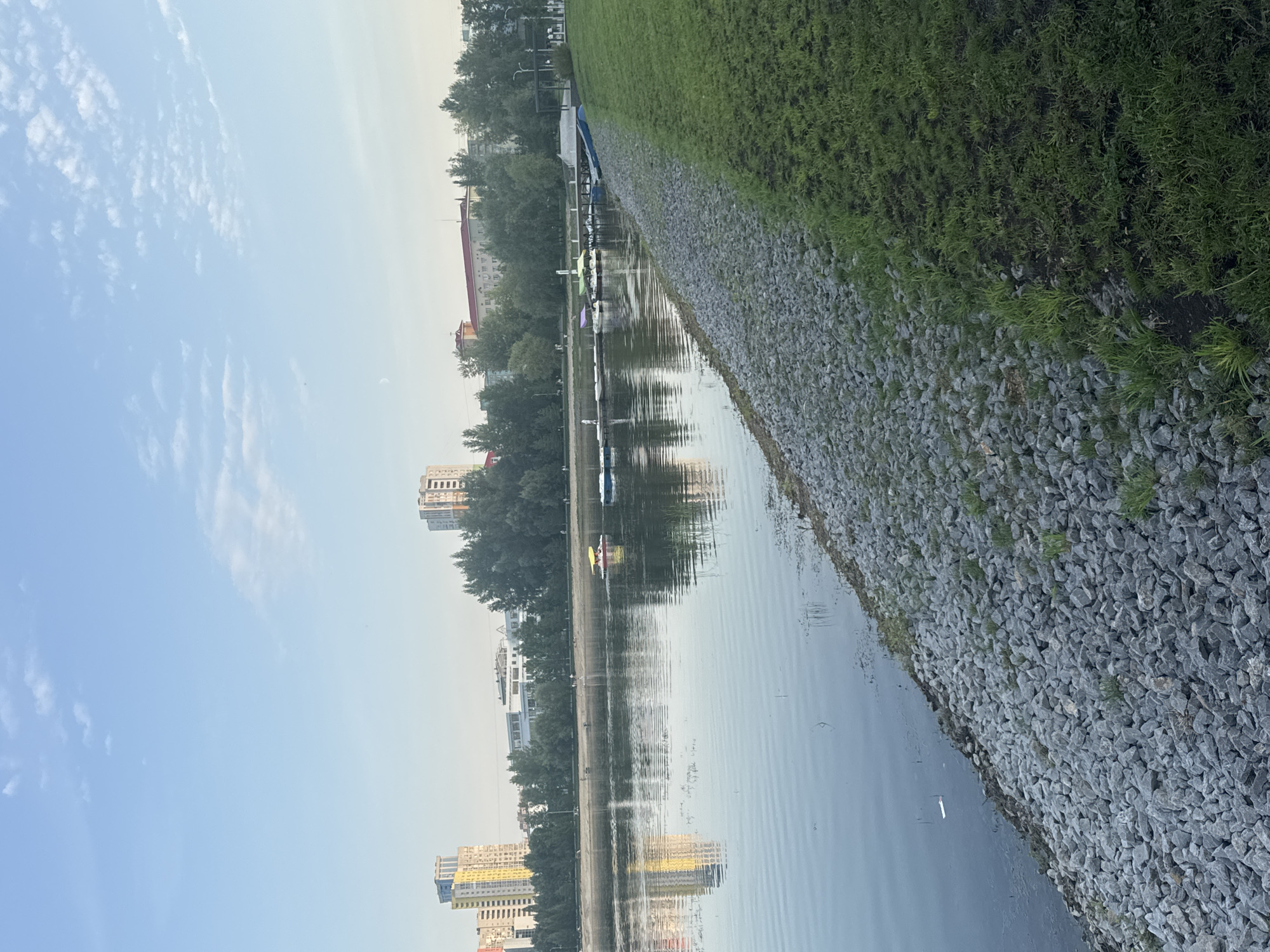 The image depicts a waterfront scene in Ufa, Russia. The foreground features a grassy embankment transitioning into a rocky shoreline. A calm body of water, likely a river or lake, occupies the left side of the frame, reflecting the sky and distant structures. Several small pedal boats are visible on the water, with one yellow boat notably out in the open. A wooden pier extends from the right side of the image, with several small boats moored to it.
In the background, a line of trees separates the waterfront from urban development. Several multi-story buildings, including tall apartment complexes with distinct architectural styles, are visible in the distance. The sky is a clear blue with scattered white clouds, suggesting pleasant weather. The overall impression is that of a public park or recreational area within the city. The presence of the buildings and the general urban environment suggest this is a developed area of Ufa.