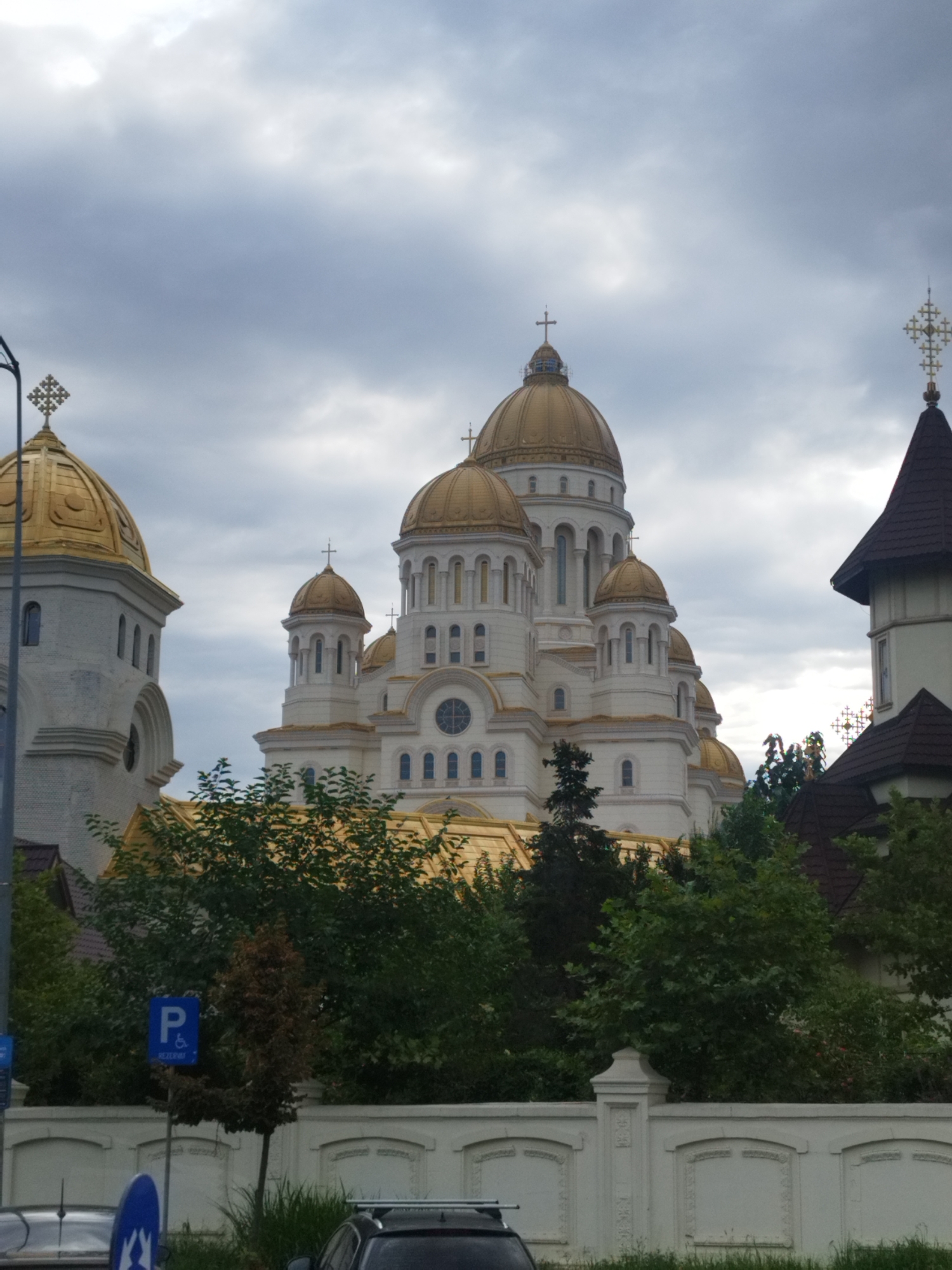 The image shows a frontal view of the People's Salvation Cathedral in Bucharest, Romania. The grand cathedral, with its multiple golden domes and white facade, is partially obscured by lush green trees and foliage. A white, decorative wall runs along the foreground, with a blue parking sign for disabled persons visible to the left of the image. The sky above is overcast with heavy, gray clouds. The scene suggests an outdoor, public space in Bucharest.
