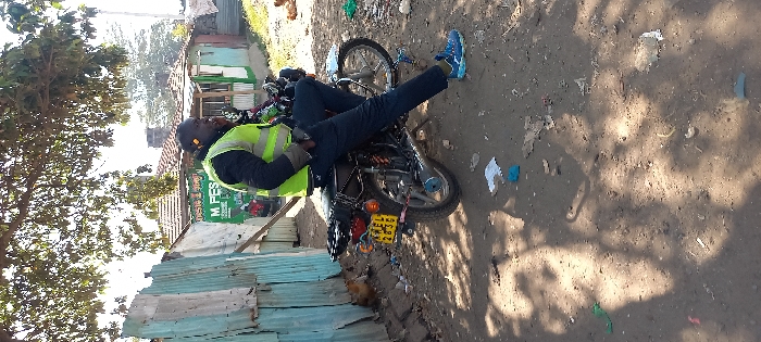 This image depicts a daytime outdoor scene in what appears to be an informal settlement or residential area of Nairobi, Kenya. In the midground, a dark-skinned male is seated on a black motorcycle, facing slightly left from the viewer's perspective. He is wearing a black long-sleeved top under a neon yellow reflective vest, dark trousers, and blue athletic shoes. His left leg is extended towards the ground, and his right hand is in his trouser pocket. He appears to be speaking into a small device held near his right ear. The motorcycle has a visible Kenyan license plate, KVEL 2B3D, affixed to its rear fender, along with a black carrier rack. To the left of the motorcycle, a fence constructed from corrugated metal sheets, some painted light blue, is visible, with a light brown chicken standing nearby. In the background, several small, single-story buildings are present; one prominently features a green 'M-PESA' sign, identifying it as a mobile money agent. This building has light blue walls and a brown tiled roof. The ground is uneven, dry dirt with scattered litter, including plastic fragments and paper. Overhead, a tree with green foliage casts dappled shadows across the scene.