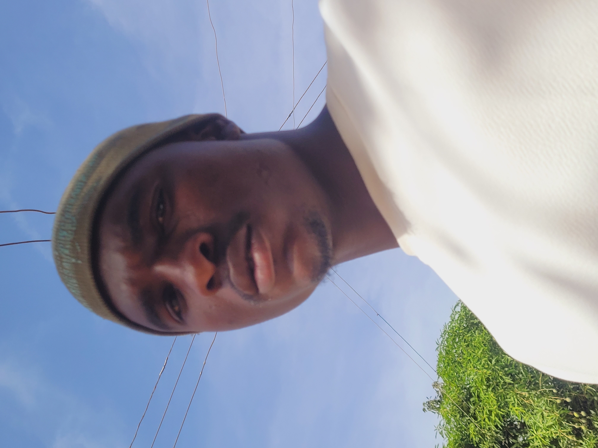 A dark-skinned male individual is depicted in a close-up, chest-up portrait, looking directly at the viewer. He wears an olive-green knitted beanie and a light-colored, possibly off-white, crew-neck garment. A short, dark goatee is visible on his chin. The background consists predominantly of a clear blue sky with minimal, diffuse white clouds. Multiple dark, thin utility wires are visible horizontally and diagonally across the upper and middle sections of the sky. In the lower-left quadrant, a dense cluster of green foliage, consistent with a tree or large bush, is present, bearing several small, light-green, oval-shaped fruits, likely mangoes. The scene is illuminated by natural daylight. This outdoor setting is located in Bukuru, Nigeria.
