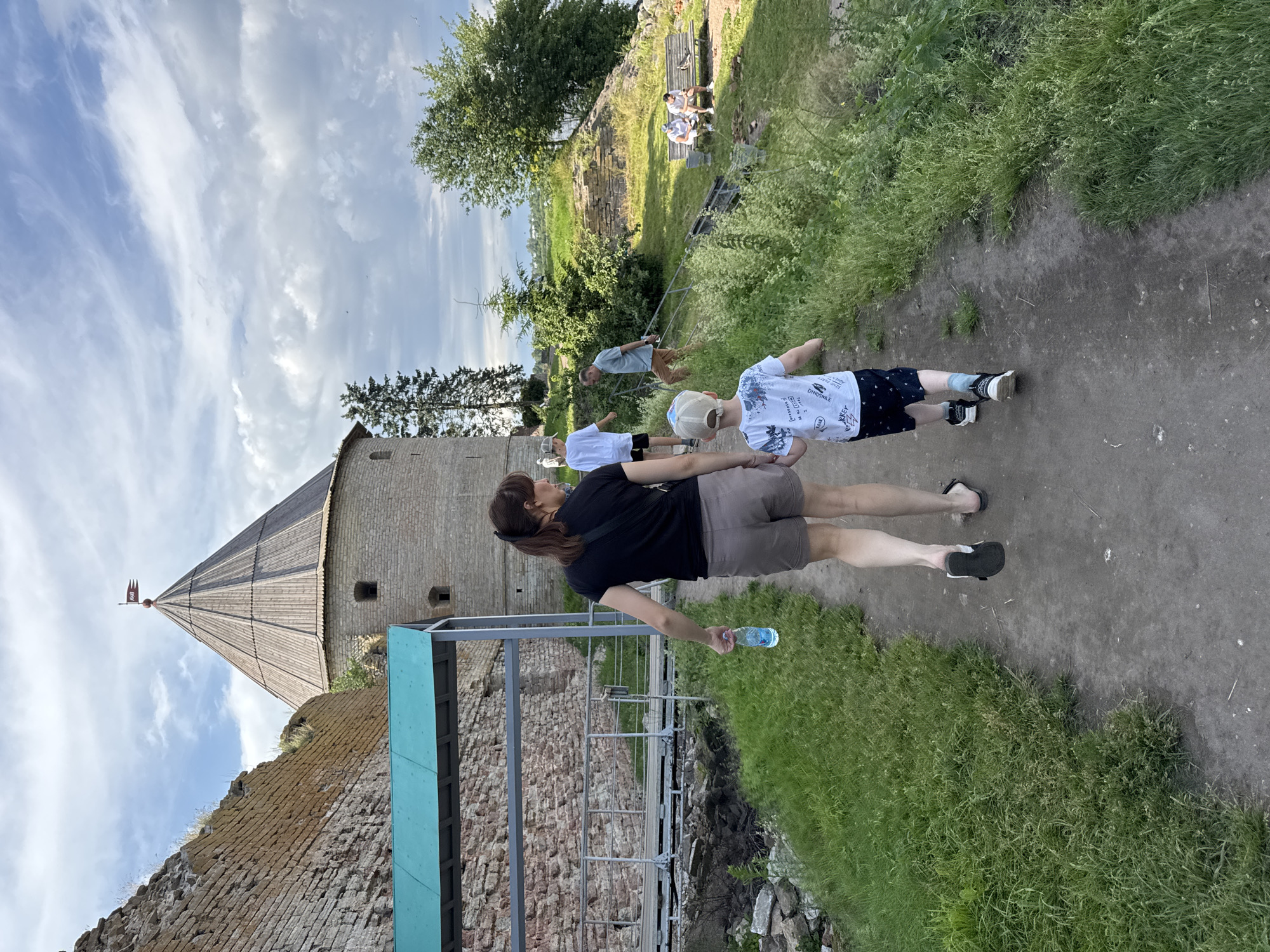 The image depicts a group of people in an outdoor historical setting, likely at the Oreshek Fortress in Shlisselburg, Russia. The primary subjects are a woman in a black t-shirt and grey shorts, holding a water bottle, and a young boy in a white t-shirt with a dinosaur print and a baseball cap. They are walking on a dirt path, with the boy in front of the woman. Several other individuals are visible in the background, some walking and others sitting on a bench.
The scene includes elements of an ancient fortress, with a prominent conical brick tower with a wooden roof and a red flag at its peak. Crumbling stone walls and overgrown grass are also visible, suggesting a well-preserved but aged historical site. The pathway is bordered by lush green grass and foliage.
The time of day appears to be daytime, with a bright, partly cloudy sky. The lighting suggests it could be late afternoon or early evening, with the sun casting some shadows. The overall mood seems relaxed and casual, possibly a tourist visit or a leisurely outing.
Visible text on the boy's t-shirt includes phrases like "DINO-SMILE," "BE READY," "BE CALM," and "BE HAPPY" in English. The water bottle held by the woman has a blue label with white lettering.