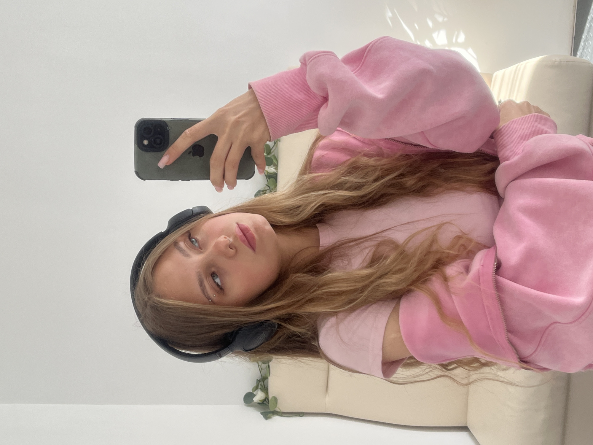 A young woman with long, light brown hair and light blue eyes is taking a selfie. She is wearing black headphones, a pink t-shirt, and an open pink hooded jacket. She has a piercing above her left eyebrow and her right hand is holding a dark green smartphone. The background appears to be an indoor setting with a white wall and a cream-colored armchair. There are some artificial green leaves with white flowers on the armchair behind her. The lighting suggests it is daytime, with sunlight casting shadows on the wall. The image was taken in the city of Фоки, Russia.