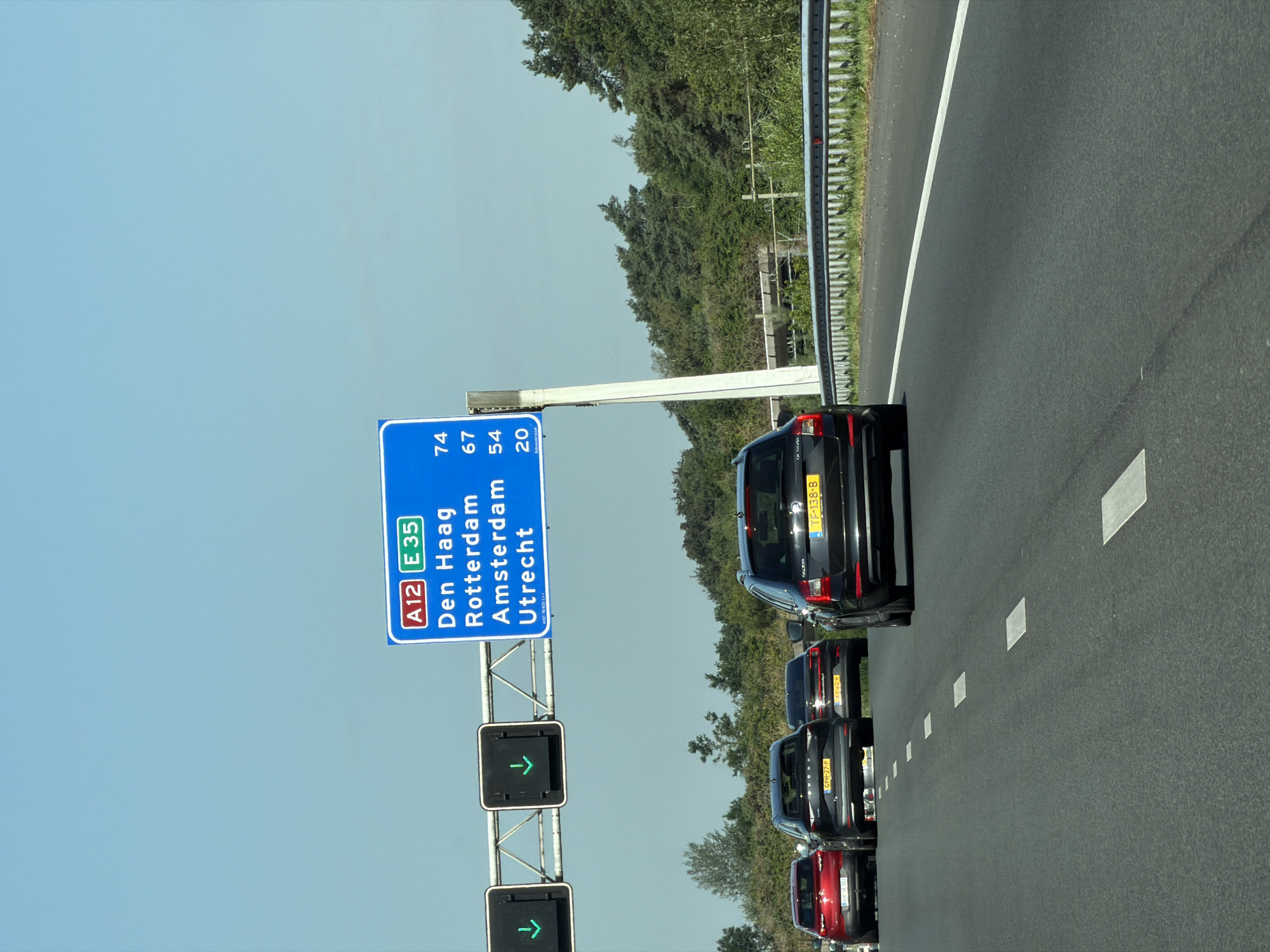 A highway overhead sign with blue background displays route information "A12" and "E 35" in red and green boxes respectively. Below the route information, a list of cities and their corresponding distances in kilometers is displayed: "Den Haag 74", "Rotterdam 67", "Amsterdam 54", and "Utrecht 20". The sign is supported by a white pole. To the left of the sign, two traffic lights with green downward arrows are visible. The scene is on a highway with several cars driving on it. The cars are a red sedan on the left, followed by two dark sedans, and a black wagon on the right. The background consists of lush green trees and a clear blue sky. The location is the A12 highway near Doorn, Netherlands.