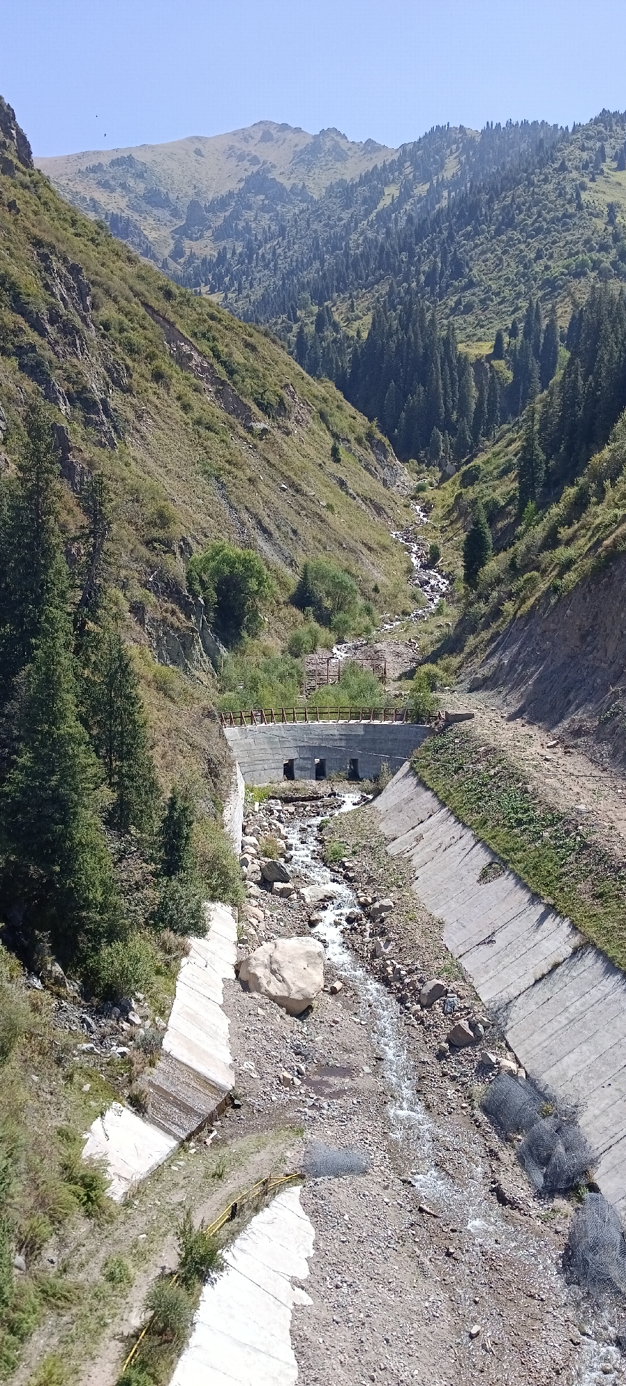 This image depicts a scenic mountain landscape with a man-made canal and a small stream running through it. The canal is constructed with concrete and appears to be designed to channel water down the mountainside. The stream is rocky and flows through the center of the canal. Lush green trees and vegetation cover the steep slopes of the mountains, which are dotted with rocks and sparse grass. The sky above is clear and blue. The scene is likely located in the mountainous region surrounding Almaty, Kazakhstan, a popular area for outdoor recreation and natural beauty.
