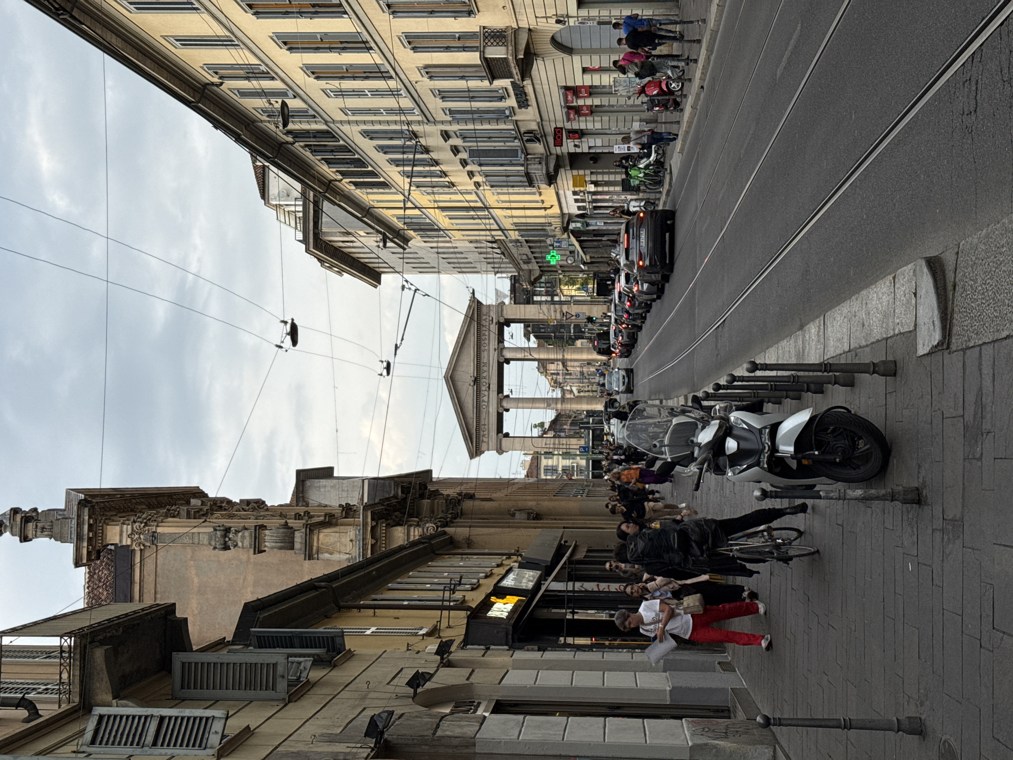 This image captures a bustling urban street scene in Milan, Italy, under an overcast sky during daytime. The perspective looks down a street flanked by multi-story buildings of classical European architecture, many featuring windows with closed shutters. Overhead wires, characteristic of tram or trolleybus systems, span across the street.
In the distance, a prominent classical archway or monument with columns and a pediment can be seen, bearing the inscription "P A C I O P V L O R V M" on its frieze, identifying it as the Arco della Pace. The street below is a mix of vehicular and pedestrian traffic. A wide asphalt road, complete with tram tracks, is occupied by a line of cars and motorcycles, including a white scooter prominently parked on the right sidewalk and a red and green motorcycle further back.
Both sidewalks are populated with numerous pedestrians, casually dressed and going about their daily activities. On the left side, a building features a brightly lit neon shop sign. A green cross sign, often indicative of a pharmacy in Italy, is visible on another building. Bollards line the edge of the sidewalk, adding to the urban infrastructure. The scene portrays a typical European city thoroughfare blending historical architecture with contemporary daily life.