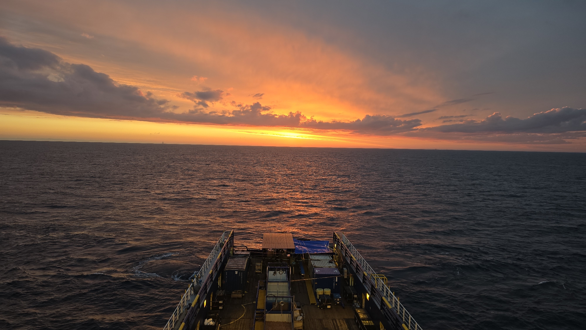 The image depicts a sunset over the ocean, viewed from the deck of a vessel. The sky is filled with shades of orange and yellow near the horizon, transitioning to darker gray clouds above. The ocean reflects the colors of the sunset, with ripples on the water's surface. The deck of the vessel is visible in the foreground, displaying containers and equipment secured with tie-down straps. The location is on the ocean, but the city and country are unknown.