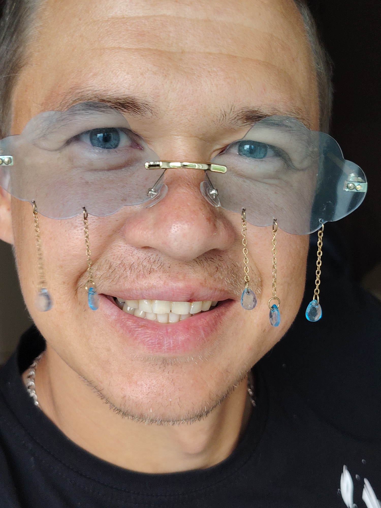 A close-up, head-and-shoulders portrait shows a person wearing novelty cloud-shaped glasses with dangling blue gem accents. The person has blue eyes and is smiling with teeth showing. They are wearing a black t-shirt and a silver chain necklace. The background is dark, suggesting an indoor setting. The image does not provide any specific geographic information, including a location in Chaikovsky, Russia.