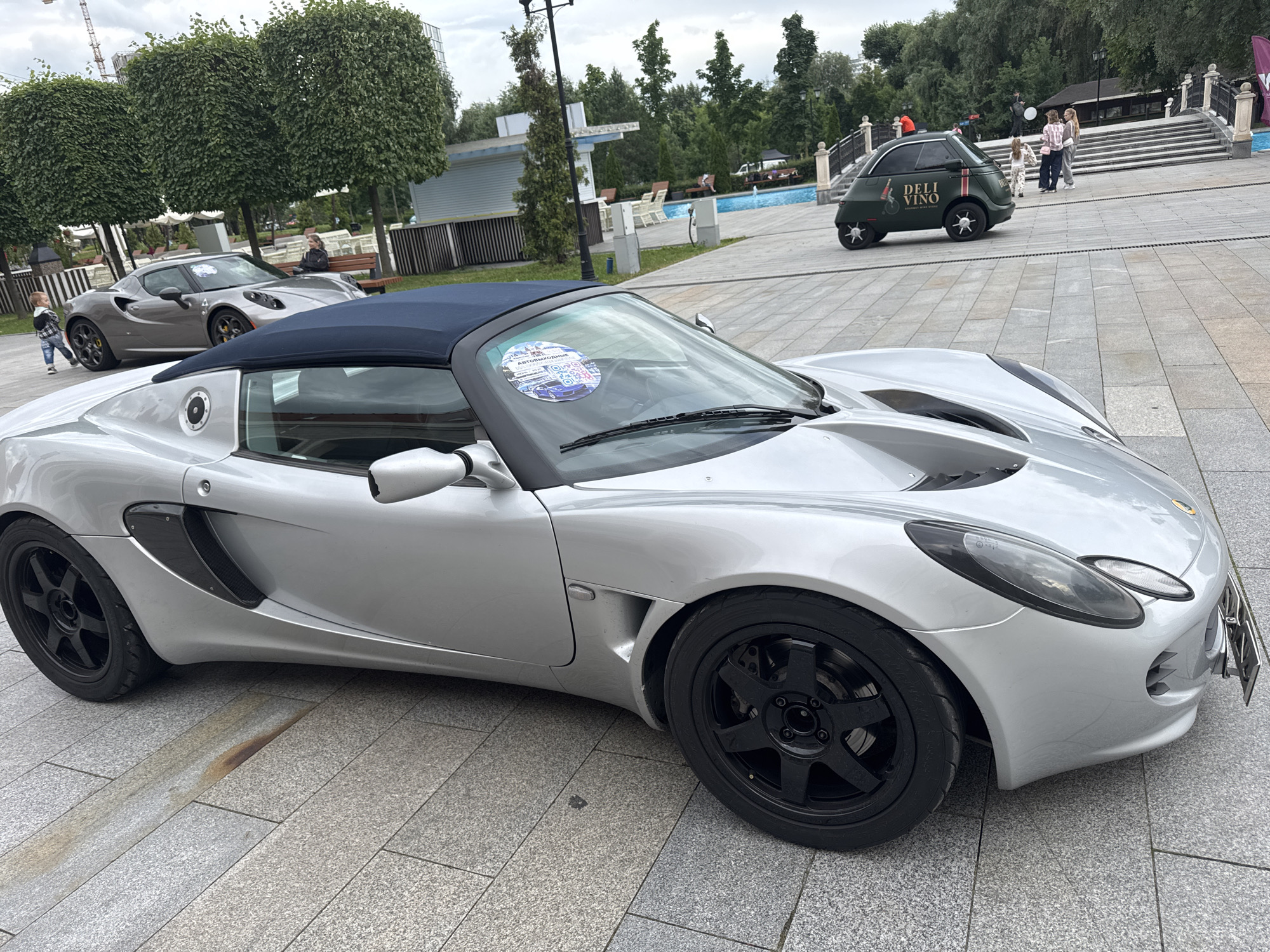 In Moscow, Russia, a silver Lotus Elise convertible is parked on a paved area, with a blue fabric top. The car has black wheels and a license plate on the front. In the background, a small green electric car with "DELI VINO" written on its side is visible, parked near a swimming pool and a set of stairs. Several people are present in the background, including a child walking near another silver car, and a group of individuals near the stairs and the green car. The environment appears to be an outdoor public space with trees and some structures.