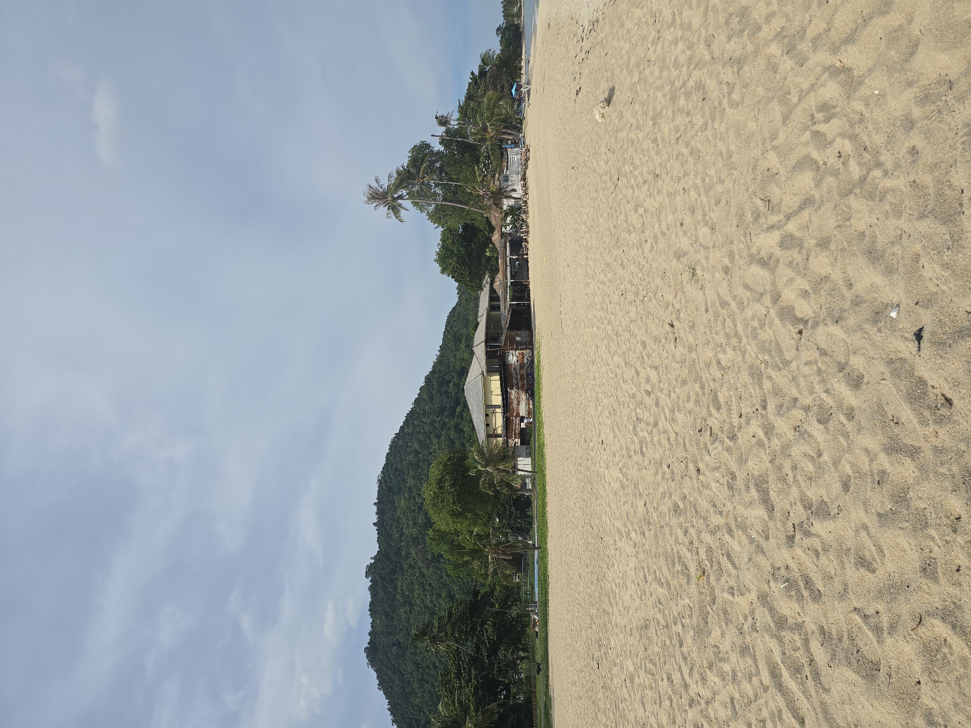The image depicts a tranquil beach scene with a sandy shore in the foreground. The sand is a light beige color, textured with ripples and footprints, suggesting it has been recently walked on. Scattered across the sand are a few small pebbles and debris.
In the mid-ground, a lush green hillside rises, covered in dense trees, including several palm trees. Nestled among the trees and at the base of the hill is a building with a yellow facade and a weathered roof, possibly a resort or guesthouse. The building appears to have multiple levels and balconies. Adjacent to the building, there are some blue lounge chairs and a swimming pool, hinting at a recreational area.
The sky above is a pale blue, with wispy white clouds. The lighting suggests it is daytime, likely late morning or early afternoon, given the brightness and the shadows cast. The overall atmosphere is peaceful and serene, evoking a sense of a tropical vacation. No people are visible in the image, and there is no discernible text. The scene appears to be a natural coastal landscape with man-made structures integrated into the environment.