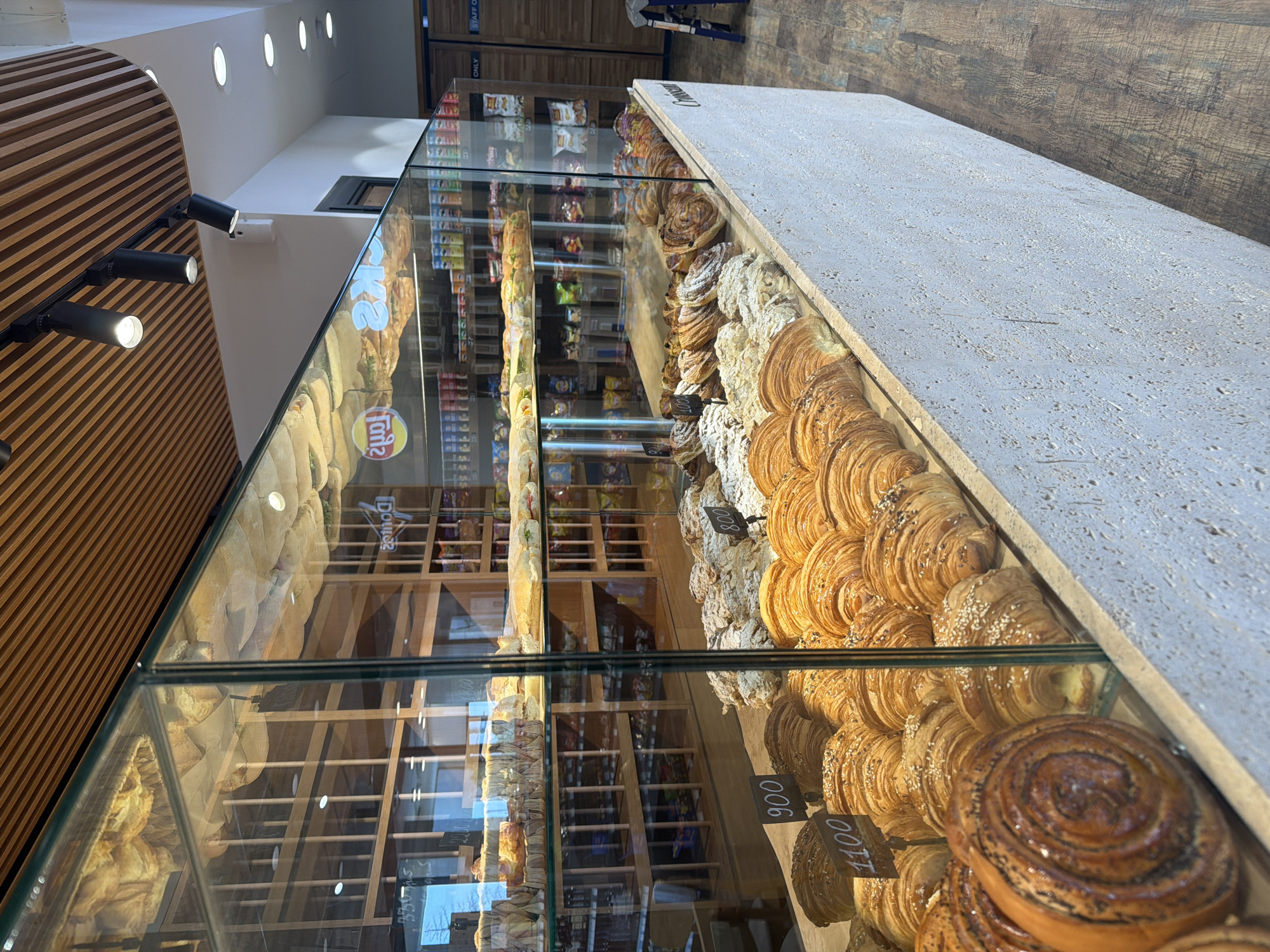 This is a forensic, matter-of-fact description of a bakery interior in Verin Ptghni, Armenia. The scene depicts a well-lit bakery with a focus on a refrigerated display case filled with various baked goods.
**Main Subjects:**
* **Baked Goods:** The primary subjects are an array of pastries and bread displayed within a glass-enclosed refrigerated case. These include numerous croissants, some sprinkled with sesame seeds and others with powdered sugar or almond slivers. There are also larger, swirl-shaped pastries, possibly cinnamon buns, prominently featured in the foreground. Several price tags with numbers such as "900", "1100", and "800" are visible among the pastries.
* **Display Case:** A large, multi-tiered glass display case dominates the foreground and mid-ground. It is constructed with clear glass panels and a light-colored, textured base that extends outwards. The interior shelves of the case are lined with paper or parchment.
* **Shelving Units:** In the background, wooden shelving units are visible, stocked with packaged goods, possibly snacks or other bakery items. Some of these packages have visible branding, including what appears to be a "Lay's" potato chip logo.
* **Ceiling and Lighting:** The ceiling features a slatted wooden design with integrated spotlights. Additional recessed circular lights are also present.
* **Floor:** The floor is covered with dark brown, wood-plank style flooring.
* **Staff Area:** In the far right background, a doorway is visible, marked with "STAFF ONLY" signage. A blue step stool is positioned near this doorway.
**Environment:**
The environment is clean and organized, suggesting a commercial establishment. The lighting is bright and functional, highlighting the products. The overall aesthetic is modern with natural wood elements.
**Actions/Interactions:**
There are no people visible in the image. The scene is static, focusing on the presentation of the baked goods.
**Objects:**
* Glass display case with refrigerated shelves
* Various pastries and bread (croissants, swirl buns, etc.)
* Price tags
* Wooden shelving units
* Packaged goods (chips, snacks)
* Wooden slatted ceiling
* Spotlights and recessed lighting
* Wood plank flooring
* "STAFF ONLY" sign
* Blue step stool
**Location Context (Verin Ptghni, Armenia):**
The presence of the bakery and the specific type of baked goods (croissants, buns) are consistent with a modern bakery offering both traditional and international items. The signage and overall presentation suggest a commercial retail space.