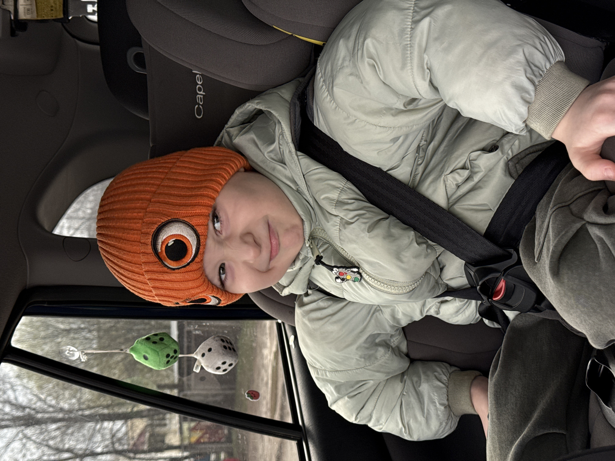 A young child is seated in a gray car seat, securely buckled in, inside a vehicle in Chaikovsky, Russia. The child, likely a boy, looks towards the camera with a gentle, contented smile. He is wearing a bright orange knit beanie with a prominent eye design on the side, and a light green puffer jacket with ribbed cuffs. A black car seatbelt crosses his chest and lap, fastened by a red and black buckle.
The car seat's headrest clearly displays the brand name "Capel". A small charm resembling a traffic light is attached to the zipper pull of the child's jacket. Through the car window to the child's left, two plush dice hang from a suction cup: one is green with black dots, and the other is white with black dots. A small, indistinct red object or sticker is also visible on the lower part of the window. The view outside the car shows bare trees against an overcast sky, indicating it is daytime during a cooler season such as winter or early spring.