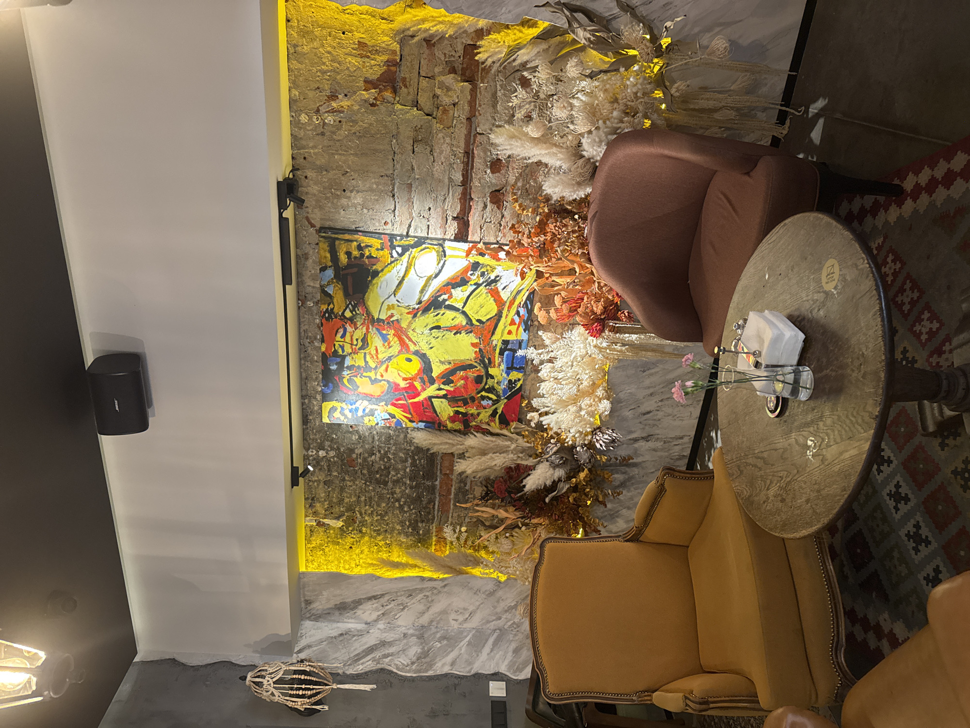 The image depicts an interior scene, likely a lounge or a cafe in an urban environment. The decor features an eclectic mix of modern and vintage elements. There are two armchairs, one upholstered in a tan or mustard-yellow fabric with decorative studs, and the other in a muted rose-brown color. Between the chairs is a small, round wooden table with a vase of flowers and napkins.
The walls are a mix of exposed brick and modern white panels, with an abstract, colorful painting mounted on the brick section. The painting appears to have a predominantly yellow color scheme with red and blue accents. The brick wall is illuminated with yellow lighting from above. Dried floral arrangements are placed around the painting and brick wall, adding a rustic touch.
The floor is partially visible, showing a patterned rug in red, brown, and gray tones. In the upper left corner, there is a black speaker mounted on the white wall. The overall atmosphere is warm and inviting.
The location is Unknown, Unknown. No people are visible. The main subjects are the furniture and decorative elements within the room. The interaction is between the objects themselves creating the design scheme and ambiance of the space.