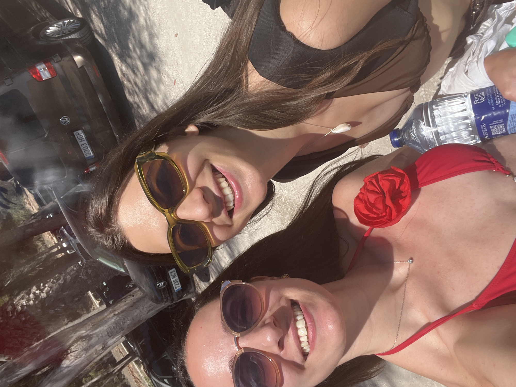 Two smiling young women in swimwear pose together in the sunshine. The woman at the top wears a black bikini top with a sheer overlay and large, amber-tinted sunglasses. The woman at the bottom wears a red bikini with a rose decoration, and rounded sunglasses with rose-tinted lenses. A bottle of Font Vella water sits nearby. In the background, the license plate "7236 NCX" is visible on one car, and "2203 HGP" is visible on another. This photo was likely taken in Teulada, Spain.