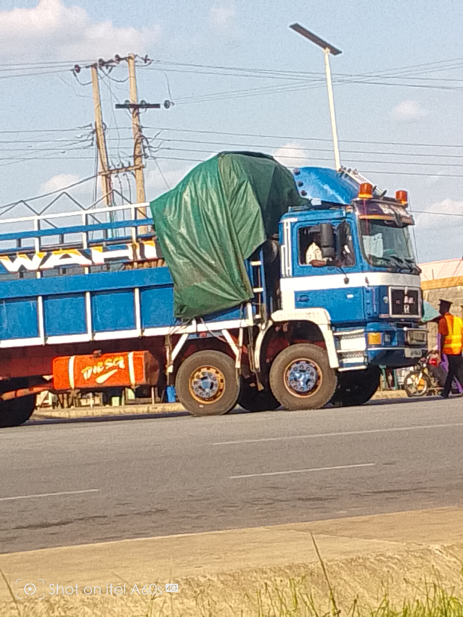 A large blue and white truck is prominently featured on a paved road in Ikot Ekpene, Nigeria, under a partly cloudy daytime sky. The truck is a cab-over-engine model with a blue cab, a horizontal white stripe, and an open cargo bed with blue railings and a white frame. A large green tarp covers a significant portion of the cargo in the truck bed. Below the truck bed, an orange fuel tank is visible, clearly labeled with the word "DIESEL" in white and red lettering. Partially visible white text resembling "WAHI" is seen on the side of the truck's cargo area. The truck has robust tires with distinctive blue hubcaps.
In the background, a network of power lines spans the sky, supported by two wooden utility poles. A tall, slender pole with a flat panel, likely a solar-powered street light, extends upwards from behind the truck's cab. On the right side of the road, a person wearing an orange safety vest and dark trousers stands near a small motorcycle. The road surface is gray asphalt with a white lane marking. A patch of dry, light-colored grass is visible in the immediate foreground. The scene depicts a typical day on an active road, possibly in a town or city. A watermark "Shot on itel A60s 4G" is present in the bottom left corner.