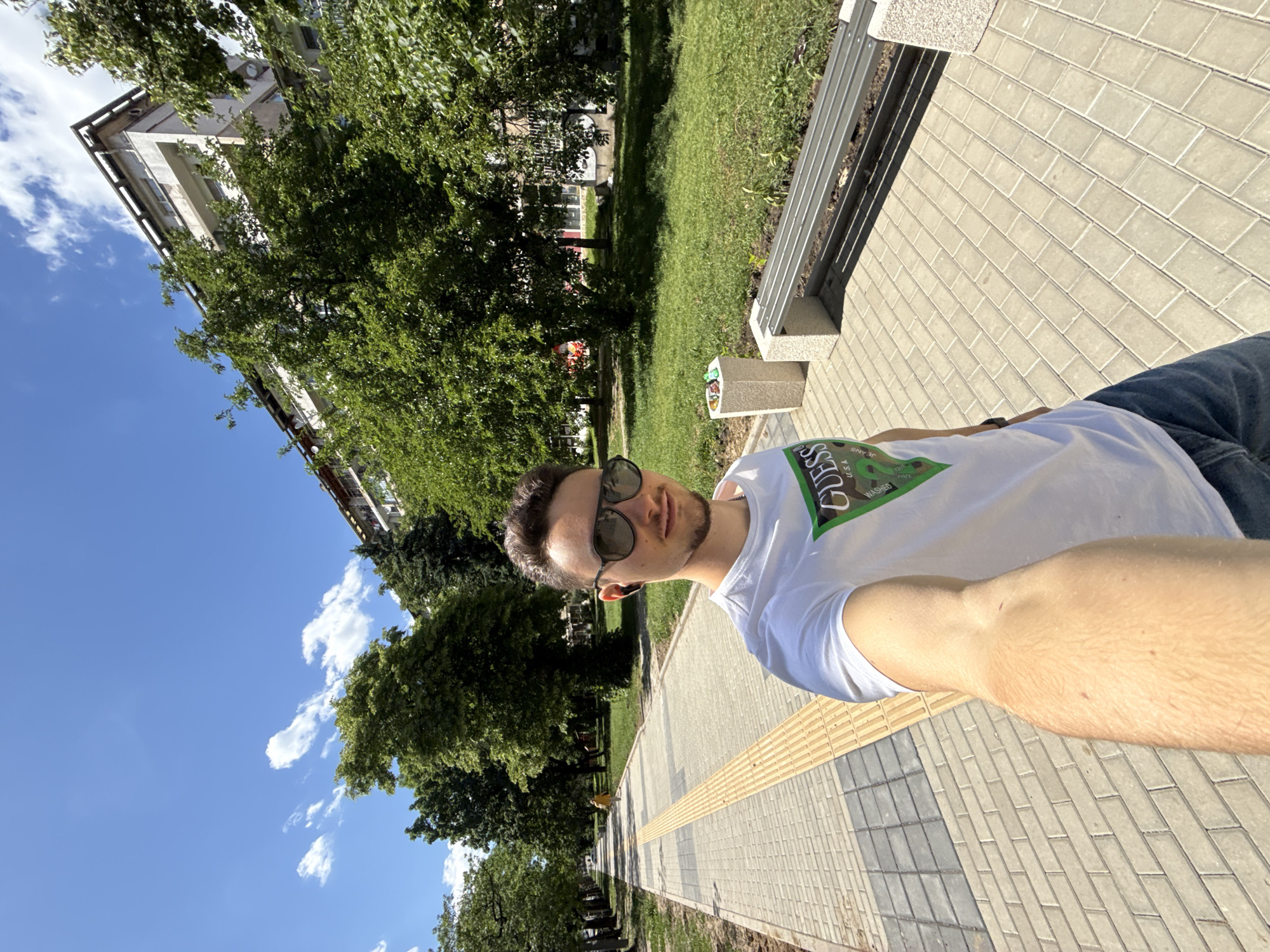 A selfie photograph, taken at a sharp angle, shows a man outdoors in Sofia, Bulgaria. The man, wearing sunglasses and a white t-shirt, appears to be standing on a paved walkway. A textured yellow strip runs along the edge of the walkway. To the left of the path are trees, grass, and a residential building. A bench sits on the other side of the path. The sky, visible behind the trees, is blue with scattered clouds.
