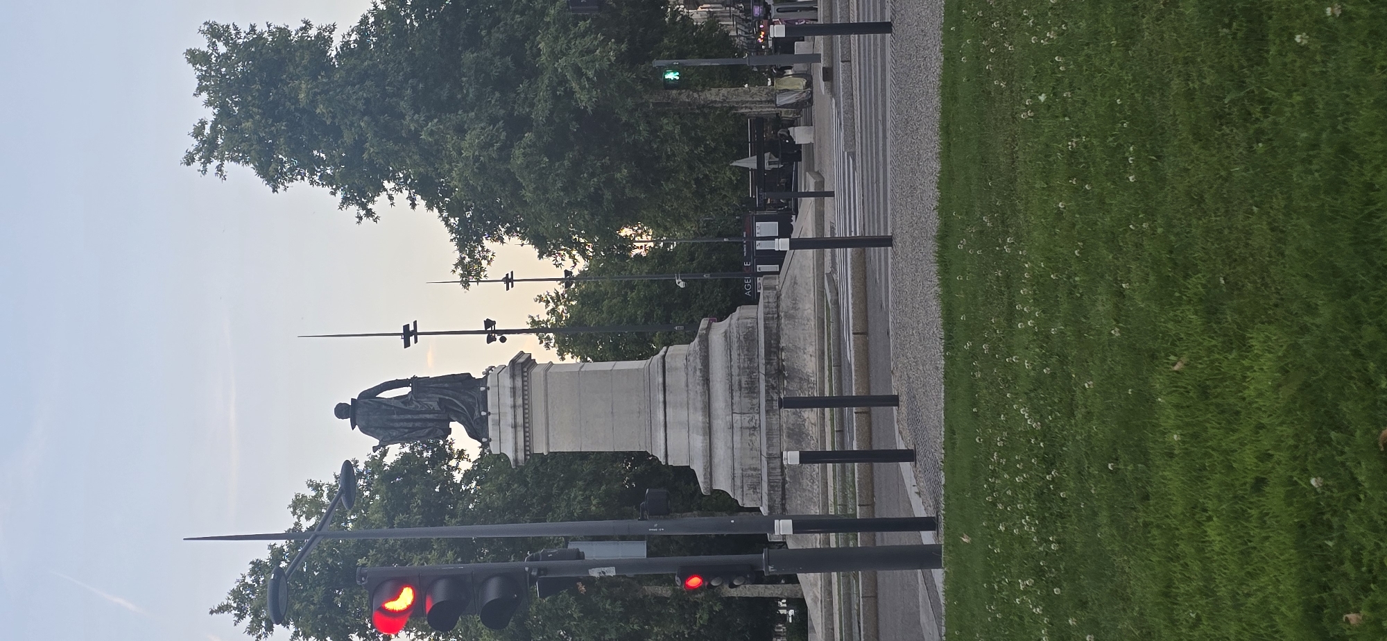 An outdoor, low-angle, full shot shows a statue of a man in a cape standing on a stone pedestal in the city of Unknown, Unknown. The statue is on the left side of the frame, and a traffic light with a red light is in the lower left. A pedestrian crossing is visible in the middle of the frame, and a green walking figure signal is visible in the distance. Trees are in the background, and a grassy area with white flowers is in the foreground. The sky is pale blue with some wispy clouds.