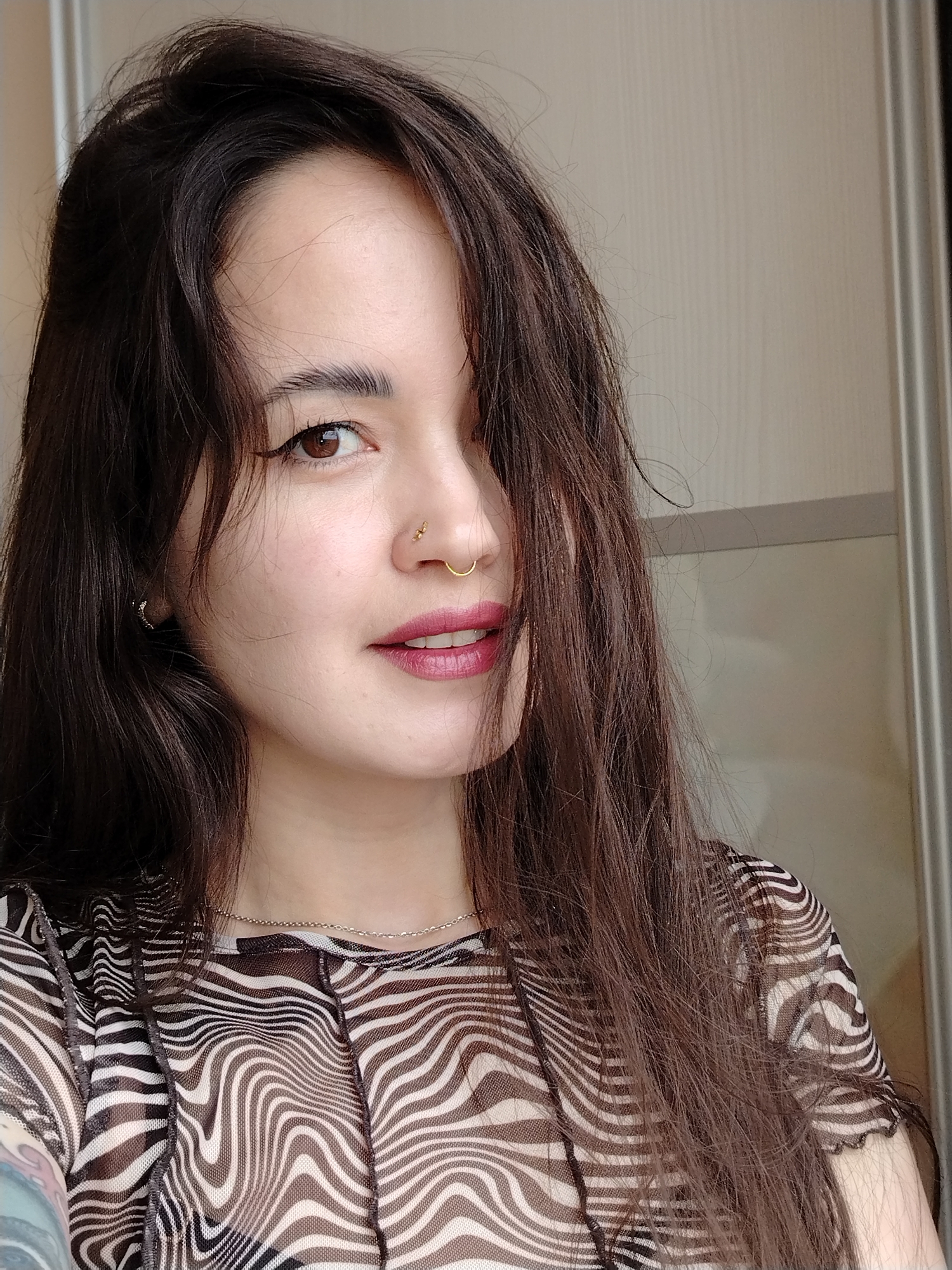 A close-up portrait features a young woman posing indoors in Noviy, Russia. Her long, dark brown hair is styled to frame her face, partially covering the right side. She has brown eyes, accentuated by winged eyeliner and defined dark eyebrows. Her lips are painted with a reddish-pink lipstick, and she offers a subtle, gentle smile, showing a hint of her teeth.
The woman wears multiple pieces of jewelry: a small gold stud on her left nostril, a gold hoop septum ring, and a delicate silver chain necklace. A small silver hoop earring is visible on her left ear. She is dressed in a sheer top with a distinctive black and white wavy, zebra-like stripe pattern. A colorful tattoo is partially visible on her left arm, peeking from under her sleeve.
The background is a simple, light-colored wall or door, suggesting an indoor setting illuminated by soft, natural light, indicating it is daytime. The woman's gaze is directed slightly off-center, connecting with the viewer. No text is visible within the image.