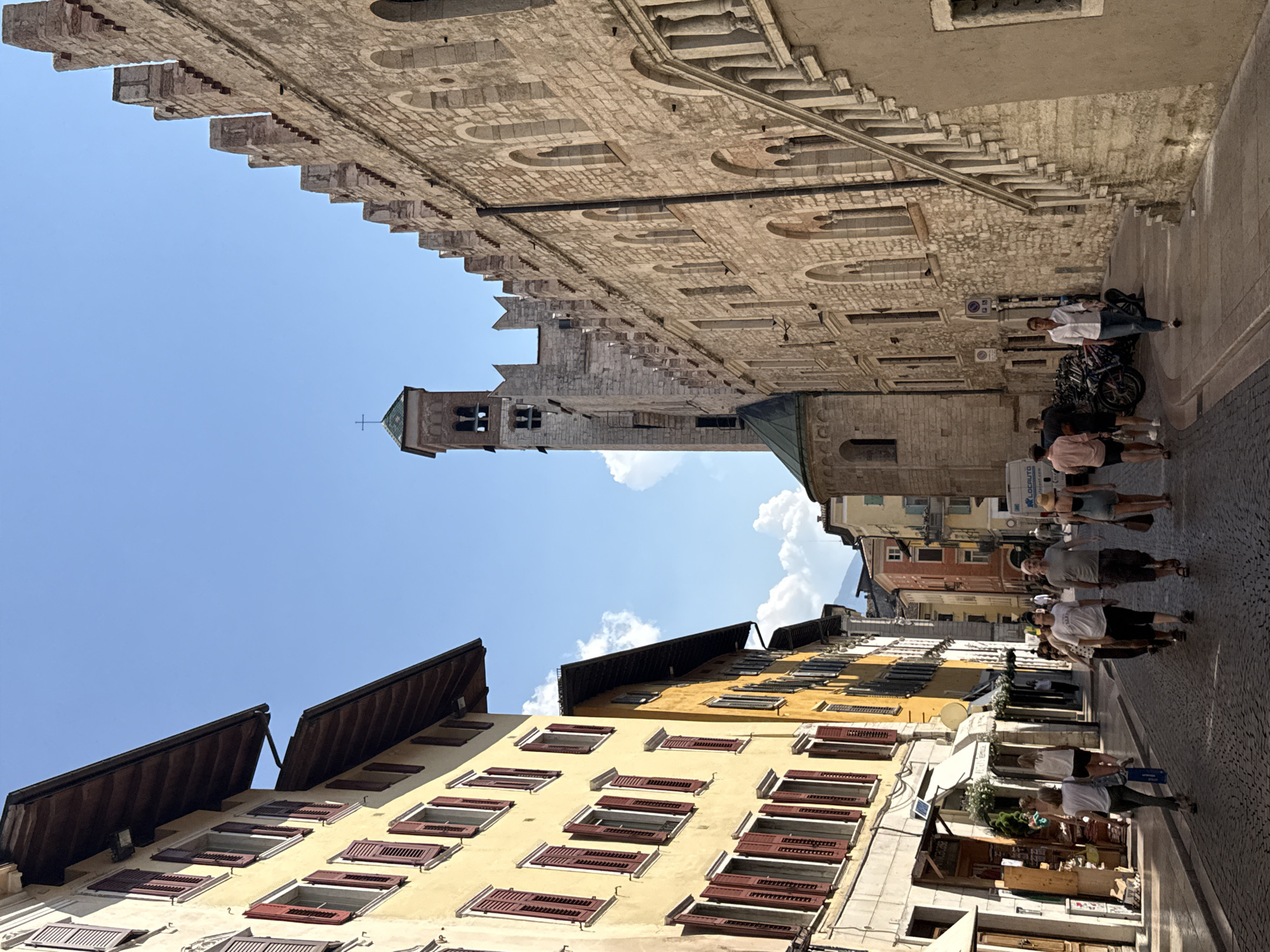 The image depicts a street scene in Trento, Italy, likely in the historic center given the architecture. On the left, a yellow building with wooden shutters lining its windows stands prominently, its pitched roof angled towards the viewer. Adjacent to it is another building, partially visible, painted a brighter yellow with similar architectural features. Shops with awnings and displays are visible at street level on the left.
The right side of the image is dominated by a large, aged stone building with a crenellated roofline, characteristic of medieval fortifications or palaces. Arched windows punctuate the stone facade, and a grand stone staircase with balustrades ascends along its exterior. Further in the background, a tall bell tower with a green roof and a cross atop it rises against the clear blue sky.
A group of people are walking down the cobblestone street. In the foreground on the left, a woman in a white top and dark pants is walking away from the camera, carrying a blue shopping bag. Behind her, a group of three men and two women are walking towards the camera. One man wears a light-colored t-shirt and dark shorts, another a dark t-shirt and shorts, and the third a light pink shirt and dark shorts. The women are dressed in casual attire. Further down the street, more pedestrians are visible, including a woman in a white shirt and blue jeans walking towards the camera, and a woman in a hat and denim shorts walking away. A van with "LOCAUTO" branding is parked on the right side of the street. Bicycles are parked along the stone wall. The overall atmosphere is that of a sunny day in a European city.