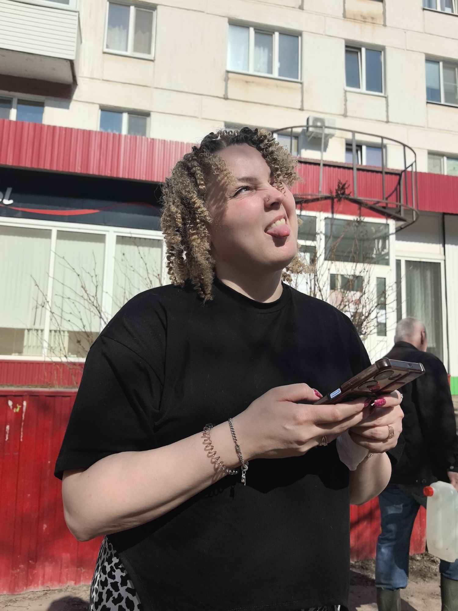 A young woman stands in the foreground, sticking her tongue out and looking upwards. She is holding a smartphone in her hands. She is wearing a black shirt and a bracelet on her left wrist. Her hair is styled in blonde curly braids.
Behind her, the facade of a multi-story building is visible. The building has a red awning over a section of the first floor, and multiple windows on the upper floors. There is a spiral staircase visible on the right side of the building. In the background, an older man is walking away from the camera, carrying a white container.
The location is likely Chaikovsky, Russia, given the prompt. The image appears to have been taken outdoors on a sunny day.