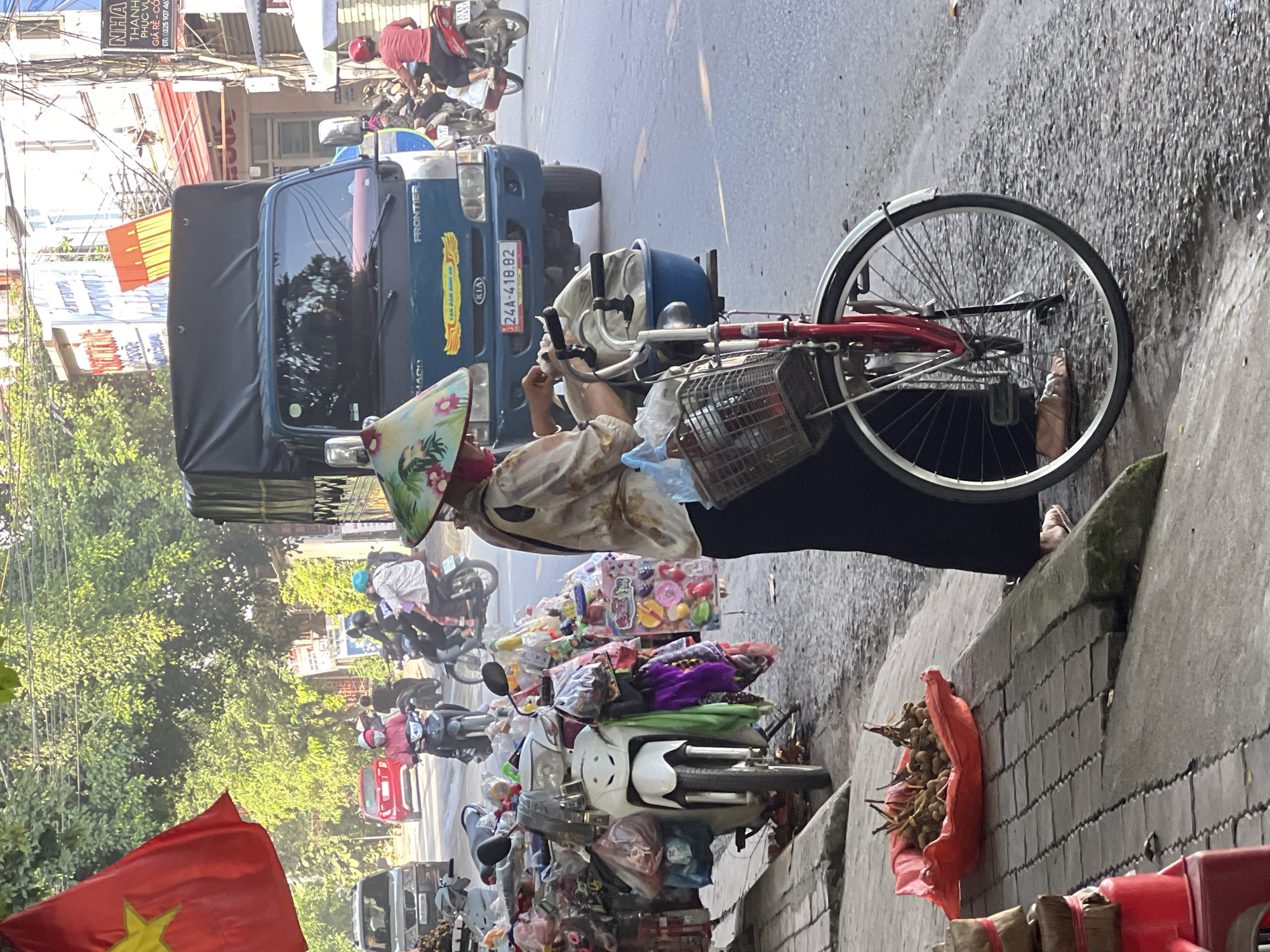 In Liên Sơn, Vietnam, a street scene unfolds with a woman, wearing a conical hat adorned with floral patterns and a patterned blouse, standing by her bicycle which is laden with a blue basin and a basket. She is actively engaged in handling some white plastic bags, possibly preparing them for sale or distribution. Her attire includes a dark skirt and sandals. In front of her, a red plastic basket overflows with a cluster of small, brown fruits, their stems still attached. To her left, a white scooter is parked, its front covered in a variety of colorful plastic toys packaged in plastic bags. The background reveals a busy street with vehicular traffic, including a blue truck with "FRONTIER" and "KIA" branding, and several motorbikes. Red and white buildings line the street, some with awnings and signage. A red and yellow Vietnamese flag flutters in the upper left corner of the frame. The ground is paved with concrete slabs and asphalt, with some debris and shadows indicating a sunny day.