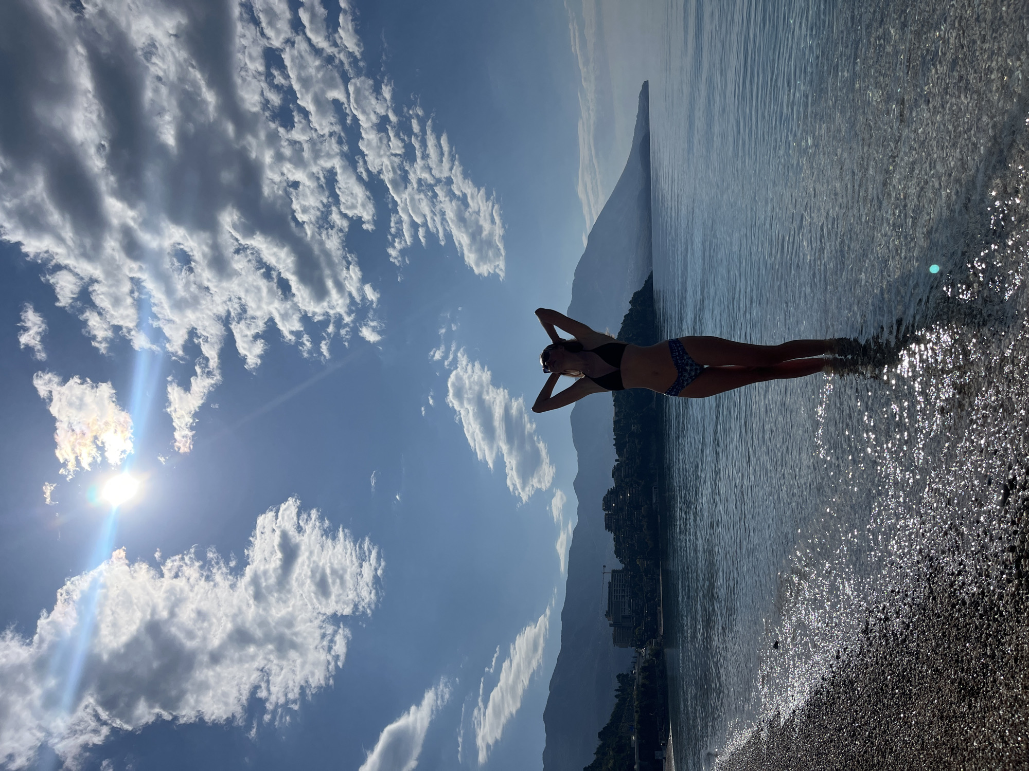 The image is a landscape scene with a woman standing in the water on a pebbled beach, with a mountainous backdrop and a partly cloudy sky. The woman is wearing a dark bikini top and patterned bikini bottoms, with her hands raised above her head. The water is clear and reflects sunlight, creating a shimmering effect. In the background, there are mountains and what appears to be the coastline of the city of Budva, Montenegro, with buildings visible along the coast. The sky is bright blue with fluffy white clouds, and the sun is shining brightly, casting a lens flare in the image. The composition suggests a tranquil, sunny day at the beach.