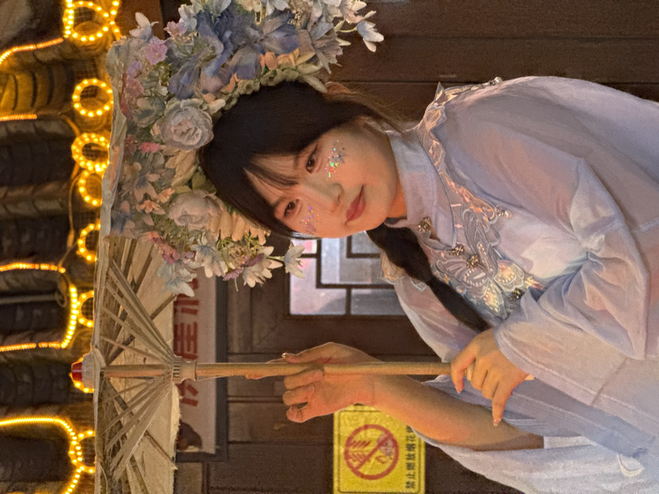A young East Asian female is depicted in a medium shot, positioned slightly to the viewer's right and looking towards the viewer's left with a gentle expression. Her dark hair is styled with bangs and a visible braid over her left shoulder. Glitter is applied to her cheeks and around her eyes. She wears a light blue, traditional Chinese-style garment featuring a high collar with ruffled trim and an embroidered or appliquéd design, possibly a butterfly, on the chest, accented with small metallic embellishments. Adorning her head is a large, ornate headpiece composed of artificial flowers in shades of blue, purple, pink, and white. She holds a light-colored, open parasol with a wooden handle upright in both hands.
The background reveals dark, traditional Chinese architectural elements illuminated by bright yellow string lights draped along the roofline and structural features. A yellow rectangular sign with a red border and Chinese characters is visible on the left, displaying a crossed-out fireworks symbol and the text "禁止燃放烟花爆竹" (No fireworks allowed). The scene is consistent with an evening or nighttime setting in a cultural or tourist area, such as Hongyadong Commercial Street in Yuzhong District, Chongqing, China.
