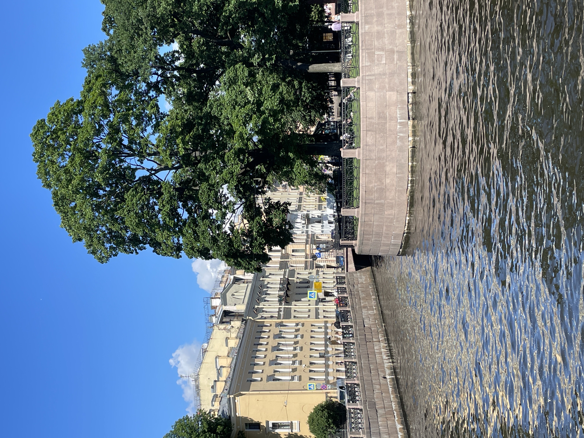 The image depicts a sunny day in Saint Petersburg, Russia, likely on the Griboyedov Canal. In the foreground, a canal with reflective, rippling water flows to the right. The canal is bordered by a stone embankment. On the left side of the canal, a row of pale yellow historic buildings with white-framed windows lines a walkway. A black metal fence separates the walkway from the canal. Signs indicating pedestrian crossings are visible along the walkway. Several cars and a few pedestrians are present on the walkway. On the right side of the canal, a circular stone structure forms a small island or point, also bordered by a black metal fence. Large, lush green trees with dense foliage dominate the right side of the image, casting shadows. Beyond the trees and the canal, more buildings are visible in the background. The sky is a clear, bright blue with a few scattered white clouds.
