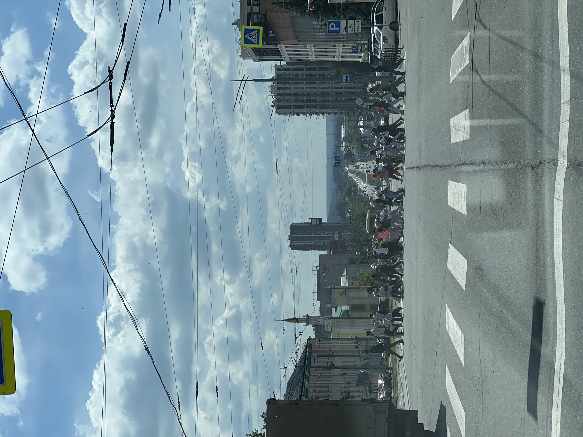 This image captures a vibrant daytime street scene in Izhevsk, Russia, with a large group of people traversing a crosswalk. The photograph is taken from a slightly elevated position, looking down the street and across the pedestrian crossing.
The primary subjects are a procession of 20-30 or more individuals, predominantly dressed in light-colored clothing and many wearing white hats or head coverings, suggesting a unified group or event. They are actively crossing a marked pedestrian crosswalk, moving from left to right across the frame. In the immediate foreground, to the left of the crossing, an individual in a dark uniform, possibly a security officer or military personnel, is also visible, appearing to be part of or overseeing the group.
The setting is an urban environment characterized by a wide asphalt road with clear white markings for the pedestrian crossing and a continuous white line along the roadside. Tall buildings line both sides of the street, including multi-story residential or commercial structures and older, more ornate architecture. A distinctive building with a golden, pointed spire or dome is visible in the mid-ground, possibly a mosque or church. Numerous overhead wires, likely for power or public transport like trolleybuses, crisscross the bright blue sky, which is dotted with large white clouds.
Visible objects include a white parked van or car on the right side of the road, and other vehicles can be seen further down the street in the distance. Several road signs are present: a yellow diamond-shaped sign with a black pedestrian symbol indicating a pedestrian crossing, and blue square parking signs marked with a white "P" and additional text. One such sign clearly displays "8ч-20ч" (8 AM - 8 PM) and directional arrows with distances like "↑ 50м" (50 meters ahead), detailing parking regulations.
The weather appears fair and sunny, indicated by the bright sky and shadows cast on the road. The overall scene depicts a busy city street with an organized public movement taking place.