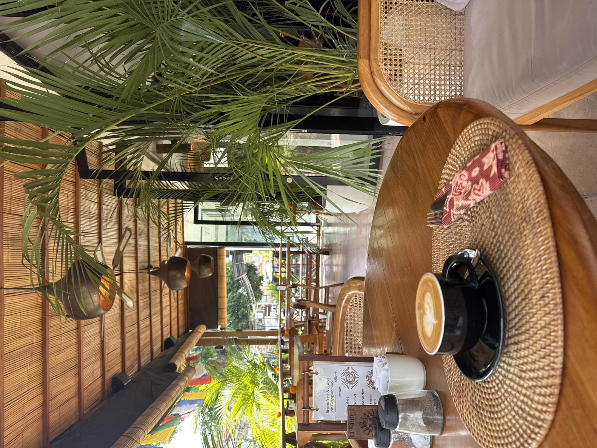 A cappuccino is seen in a black cup on a round wooden table in a restaurant called La Kantina, which serves breakfast and juice until 12 pm. The table also has a woven placemat, a red napkin, a fork, a menu, sugar, and a salt shaker. The restaurant has a bamboo roof and open walls, with palm trees and other plants visible outside. Other wooden tables and chairs can be seen in the distance. The image was taken from the perspective of someone sitting at the table, in Unknown, Unknown.