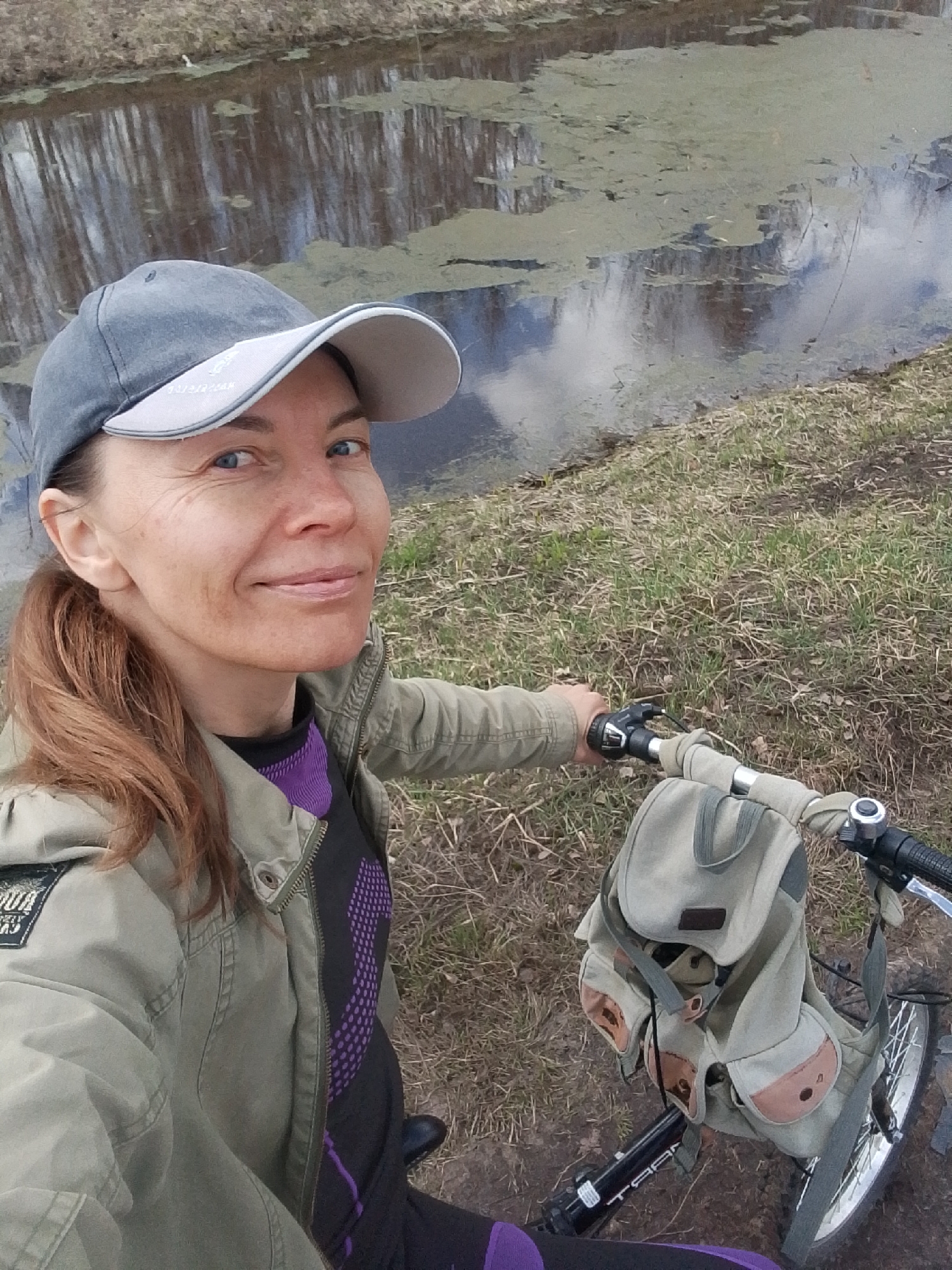 A woman with reddish-brown hair pulled back in a ponytail is pictured in a selfie while on a bicycle ride in Снопок Новый, Russia. She is wearing a grey baseball cap, a black and purple patterned shirt, and a light olive-green jacket. She has a slight smile and is looking into the camera. The bicycle has a khaki-colored bag attached to the handlebars.
In the background, there is a body of water, possibly a canal or pond, with reflections of trees and sky. The water's surface has some green algae or vegetation floating on it. The ground near the water's edge and bicycle is covered with grass and dry vegetation. The setting appears to be a park or natural area within or near Снопок Новый.