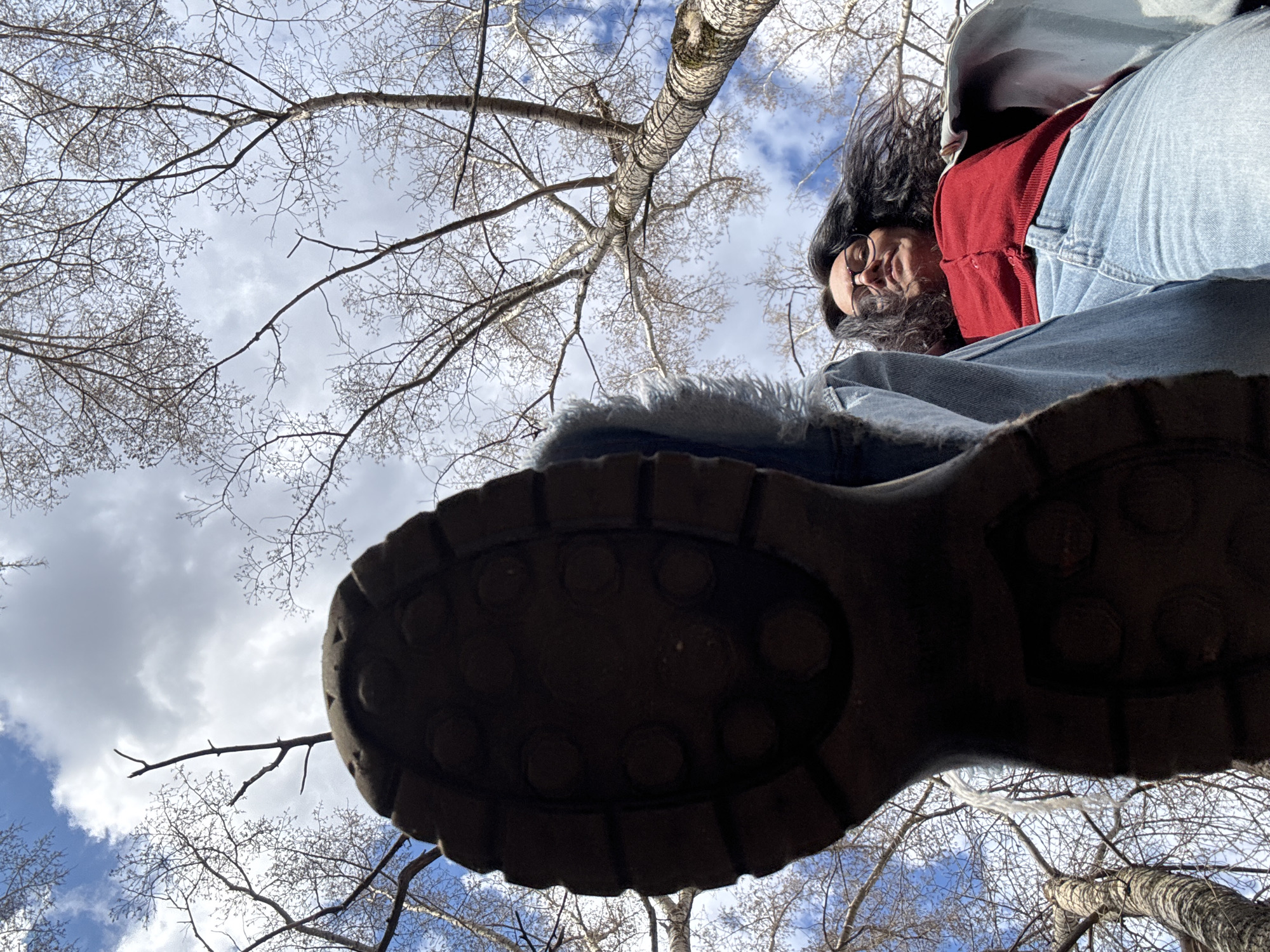 The image presents a unique low-angle perspective, looking upward from the ground. The most prominent subject in the foreground is the dark, textured sole of a large boot, suggesting a hiking or work boot, positioned directly above the camera. Above and to the right of the boot, a person is visible, looking down towards the camera with a slight smile or relaxed expression. This individual has dark hair, wears glasses, a red top, and blue jeans, with what appears to be a light-colored jacket or fur-lined collar.
The background is dominated by a clear sky, featuring a mix of white and grey clouds alongside patches of blue. Numerous bare tree branches extend across the frame, forming a natural canopy. One tree trunk with light-colored bark is distinctly visible, possibly indicating a birch tree, characteristic of temperate regions.
The scene takes place outdoors during the daytime, under mixed sunny and cloudy conditions. The bare branches suggest it is late autumn, winter, or early spring. The unusual perspective creates an immersive experience, positioning the viewer directly beneath the person's foot, hinting at a playful or observational moment in a wooded or forested area. Given the location context, this natural setting could be in Chaikovsky, Russia. There is no visible text in the image.