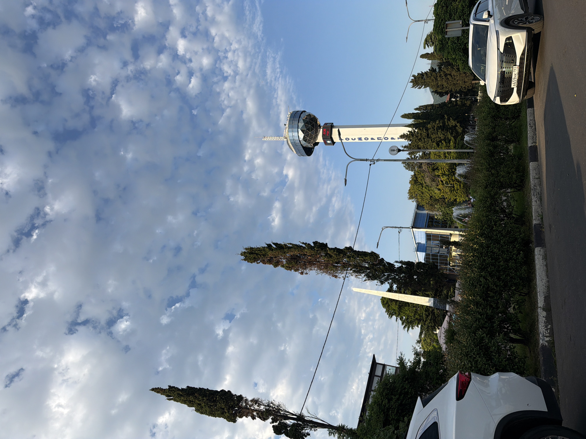 The image depicts a clear day with scattered clouds in a blue sky. The primary subject is a tall, white tower with a distinctive glass-domed observation deck at its summit. The tower has text written vertically on its side that reads "КОСМОНАВТОВ" (Kosmonavtov). Attached to the tower is a digital display showing a temperature of -9.8°C and a time of 09:19.
In the foreground, a white SUV is parked on the right side of a paved road. The car has a license plate with the text "A659KK 193". Adjacent to the road is a sidewalk and a grassy area with several tall, slender trees and lush green bushes. In the background, behind the tower and trees, a modern building with blue-tinted windows is visible, along with other greenery and distant hills or mountains.
The scene appears to be an outdoor public space, possibly a park or an area near an attraction, given the presence of the tower, fountain, and the general landscaping. The time of day is morning, as indicated by the digital display. The weather is pleasant, with a clear sky and visible clouds. The overall mood conveyed is calm and serene.