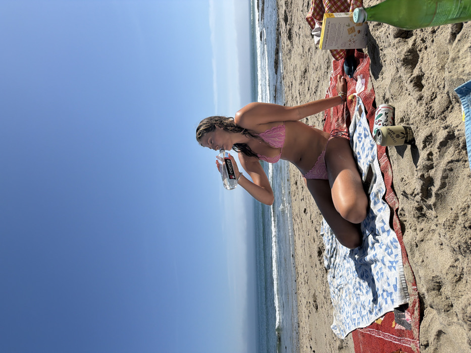 A female individual is seated on a sandy beach in Malibu, United States of America. She is observed in profile, facing left, wearing a pink lace two-piece bikini. Her hair is wet, and her skin exhibits a tan. She is actively drinking from a clear plastic bottle labeled "essentia," held to her mouth with her right hand; her head is tilted back. She rests upon two layered beach towels: a red patterned towel beneath a blue and white geometric patterned towel. To her right on the sand, a metallic can labeled "LPFND BEER," a yellow rectangular box displaying "what good food can do," a portion of a red and white checkered fabric, a dark smartphone, and a green glass bottle are visible. The background consists of the Pacific Ocean with white-capped waves breaking on the shore, extending to a distant horizon partially obscured by atmospheric haze, all beneath a clear, bright blue sky.
