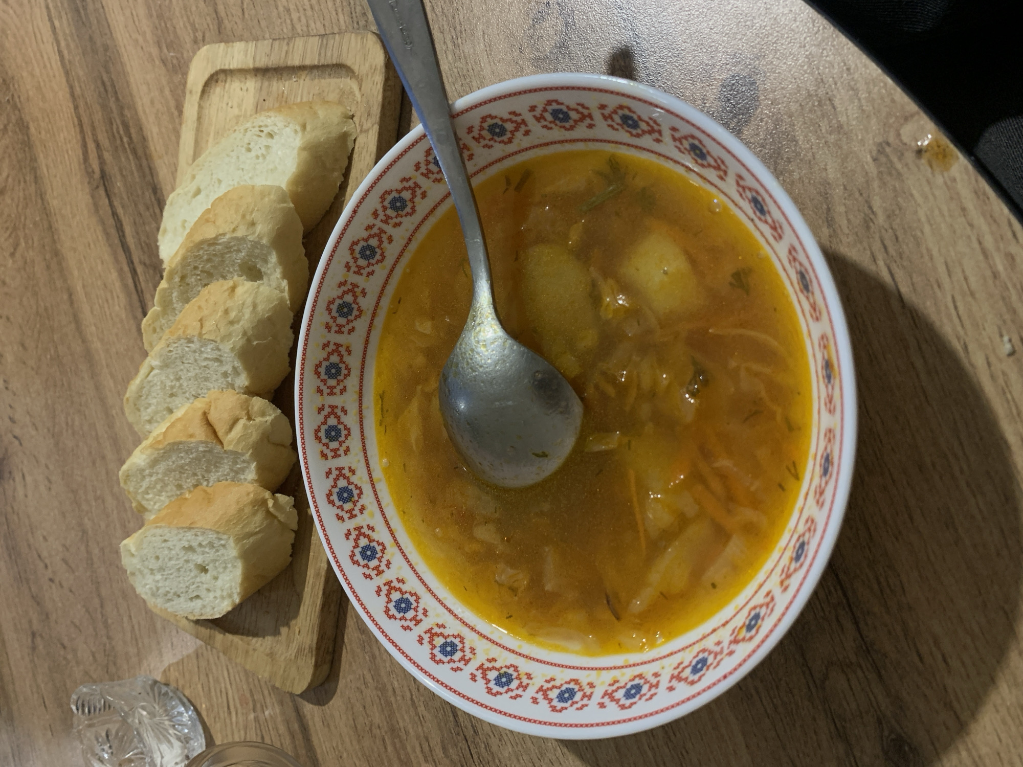 A close-up, overhead shot shows a bowl of golden-orange soup filled with visible chunks of vegetables, such as potatoes and carrots, and a silver spoon resting inside. The soup bowl has a white exterior adorned with a repeating red and blue folk-art pattern. Next to the bowl, on a light-colored wooden cutting board, are five slices of white bread arranged in a row. The food is placed on a wooden table with a visible grain pattern. In the upper left corner of the image, a small portion of a clear, cut-glass bowl is visible. The scene suggests a meal setting in an indoor environment, likely a home or restaurant, but the specific city and location are not discernible from the image.
